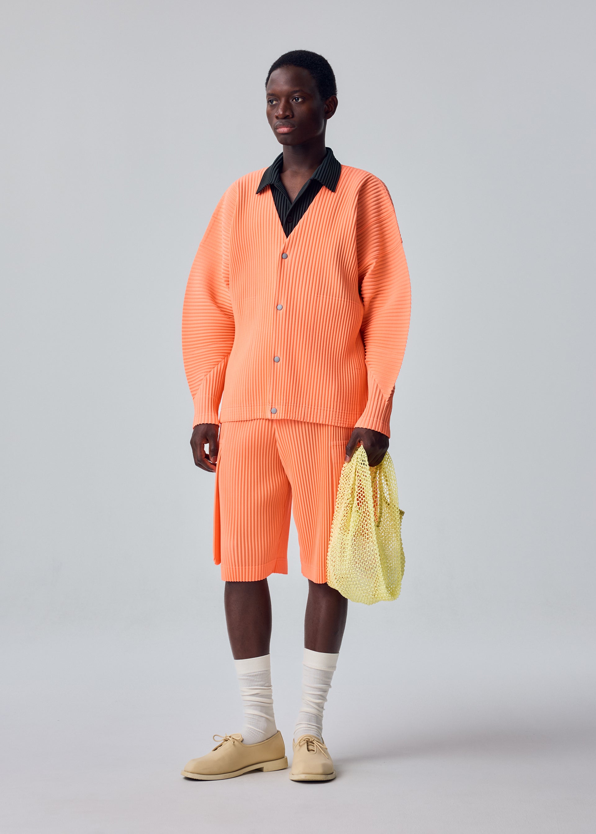 Zoomed view A person stands against a plain light background, showcasing a minimalist look featuring an orange pleated polyester cardigan and matching shorts, a black collared shirt, beige shoes with white socks, and carrying the MESH PLEATS BAG by HOMME PLISSÉ ISSEY MIYAKE.