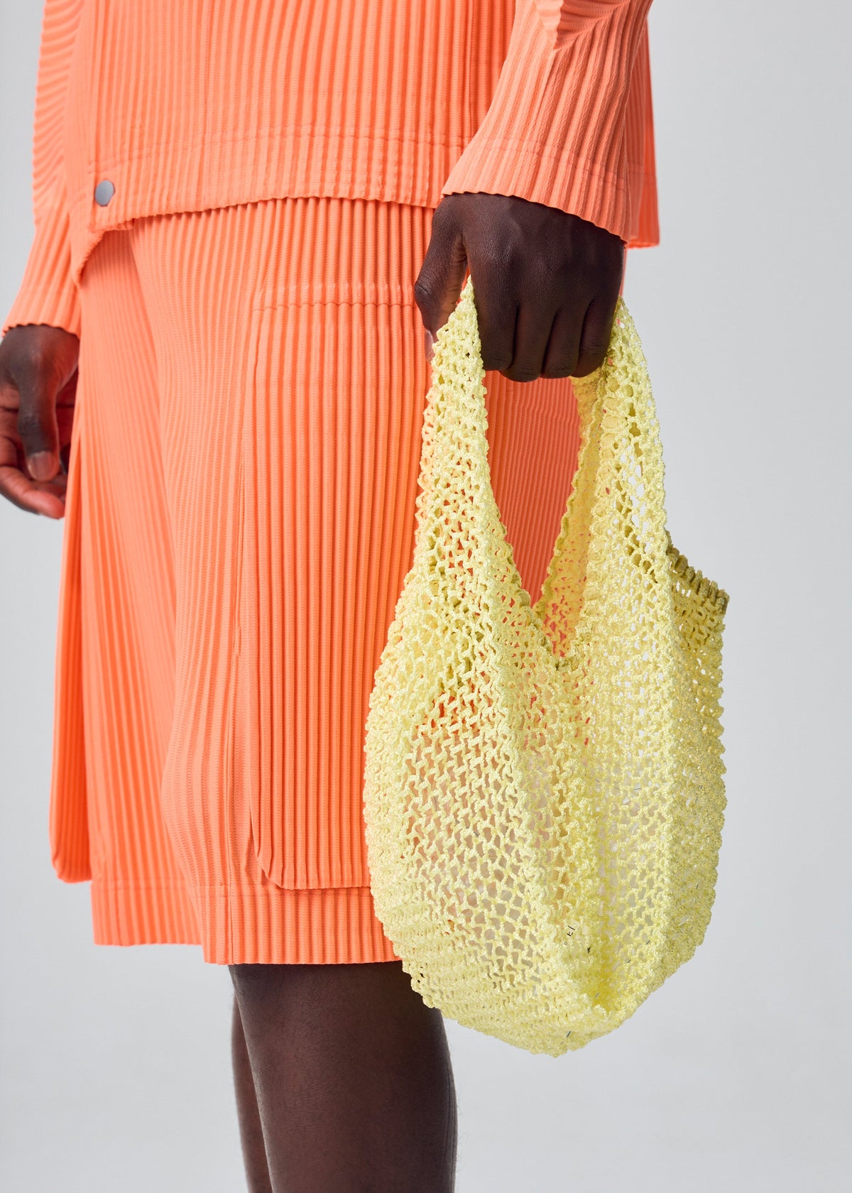 A person dressed in ribbed orange attire holds the small, yellow MESH PLEATS BAG from HOMME PLISSÉ ISSEY MIYAKE. The minimalist photo is cropped to highlight only the lower torso and hand, set against a plain background. view 7 of 7