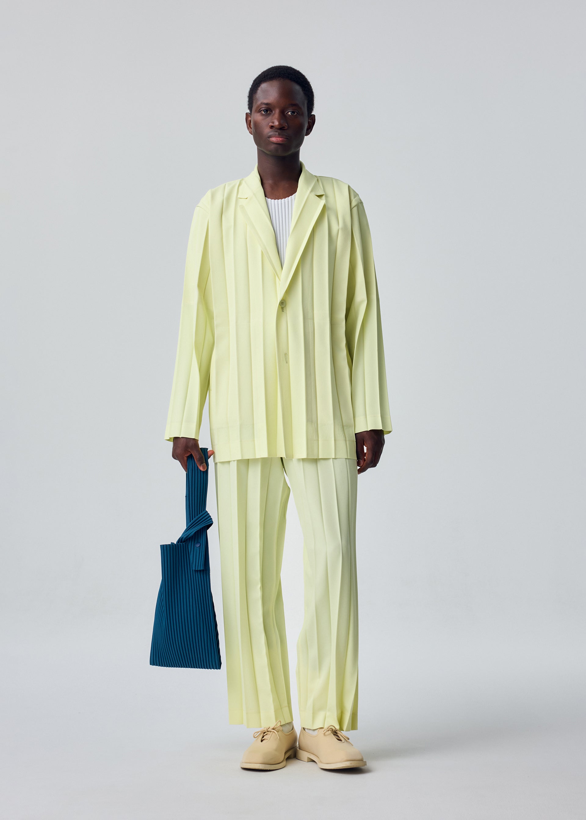 Zoomed view A person stands against a plain light gray background, dressed in a light yellow suit, white shirt, beige shoes, and holding the HOMME PLISSÉ ISSEY MIYAKE UMBRELLA PLEATS BAG in blue.