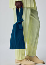 A person in light yellow pleated pants and cream shoes holds the UMBRELLA PLEATS BAG by HOMME PLISSÉ ISSEY MIYAKE—a pleated blue handbag. The photo focuses on the lower body against a plain background.