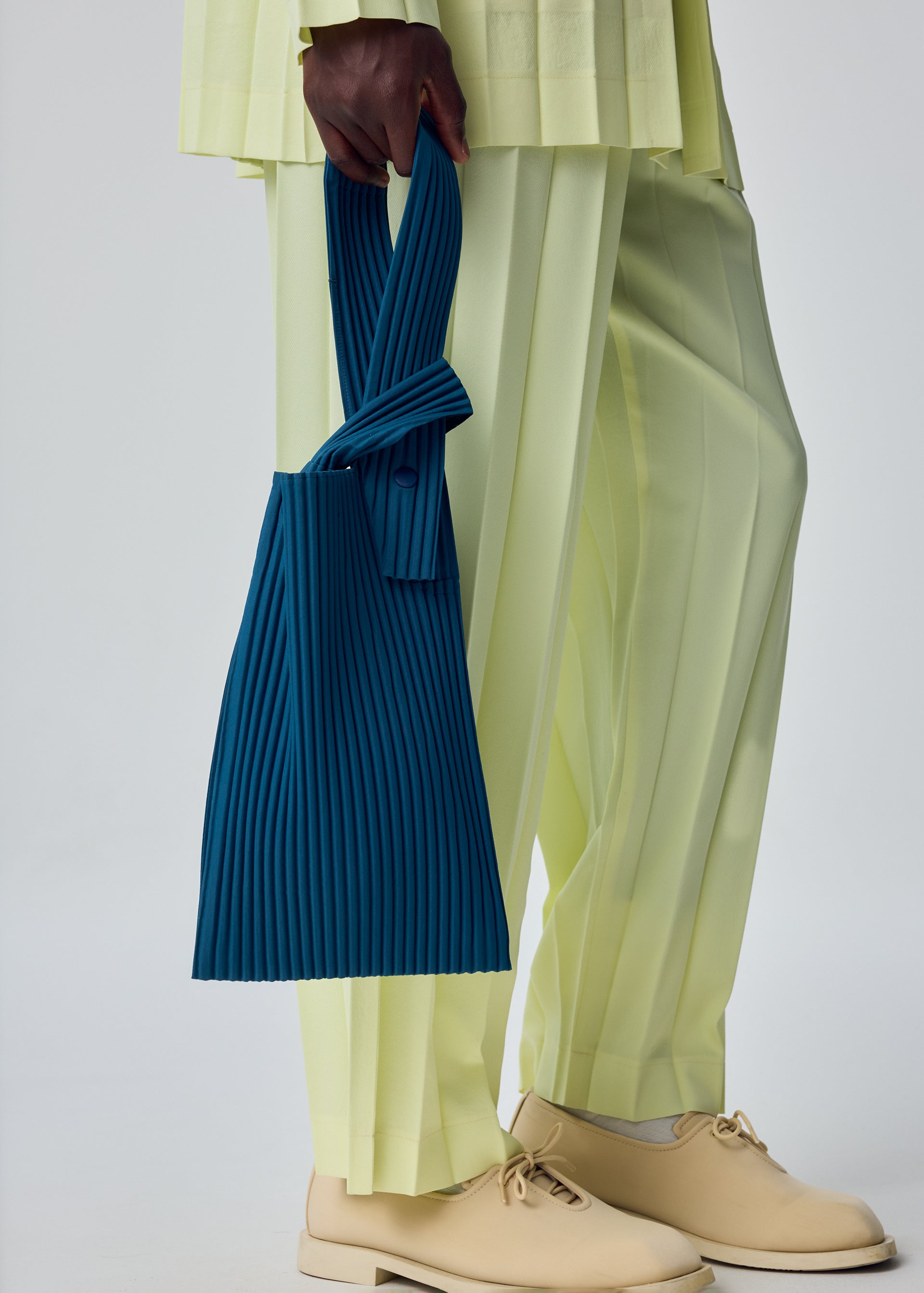 Zoomed view A person in light yellow pleated pants and cream shoes holds the UMBRELLA PLEATS BAG by HOMME PLISSÉ ISSEY MIYAKE—a pleated blue handbag. The photo focuses on the lower body against a plain background.