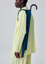 A person dressed in a light yellow pleated outfit from HOMME PLISSÉ ISSEY MIYAKE stands in profile, carrying a closed navy blue umbrella secured with the brand’s UMBRELLA PLEATS BAG across their back. The background is plain and light-colored.
