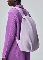 A person wearing a textured, long lavender coat carries the HOMME PLISSÉ ISSEY MIYAKE PLEATS BACKPACK with a laptop sleeve, shown from the side against a plain light gray background.