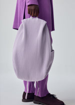 A person in ribbed lavender and purple attire holds the HOMME PLISSÉ ISSEY MIYAKE PLEATS BACKPACK with side pockets. Only their lower body and hand are shown against a plain, light backdrop.
