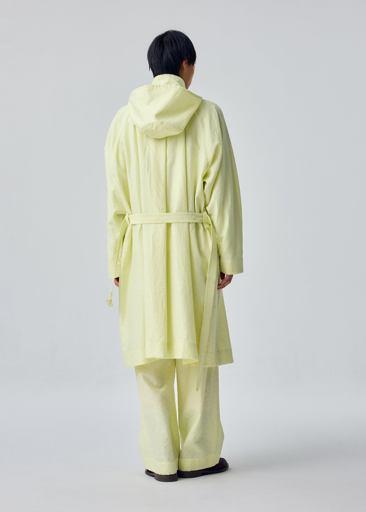 A person stands with their back to the camera, dressed in the OPACITY light yellow hooded coat and matching loose pants from HOMME PLISSÉ ISSEY MIYAKE, crafted from a nylon linen blend, set against a plain light background. view 6 of 9