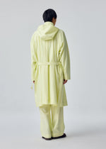 A person stands with their back to the camera, dressed in the OPACITY light yellow hooded coat and matching loose pants from HOMME PLISSÉ ISSEY MIYAKE, crafted from a nylon linen blend, set against a plain light background.