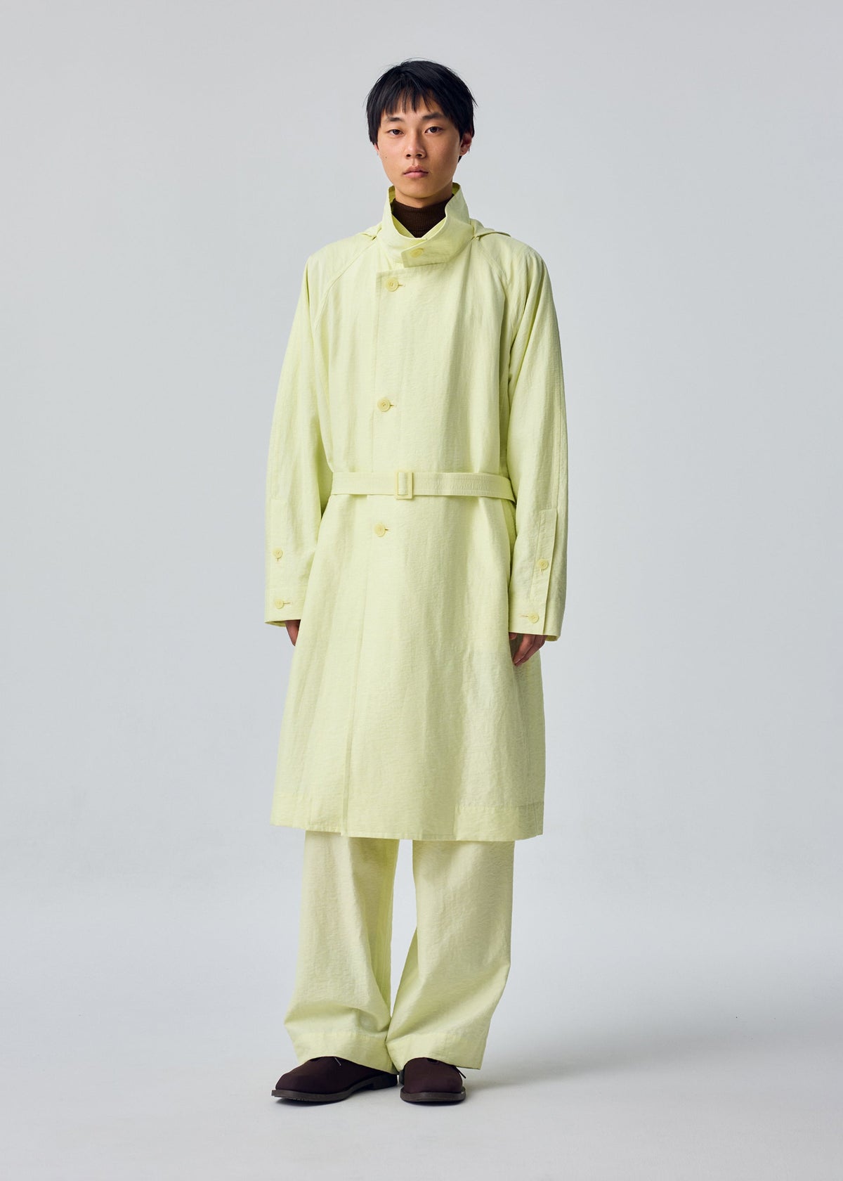 A person is wearing the OPACITY light yellow belted trench coat with a detachable hood and matching wide-leg pants from HOMME PLISSÉ ISSEY MIYAKE, styled with dark shoes and posed against a plain light background. view 7 of 9