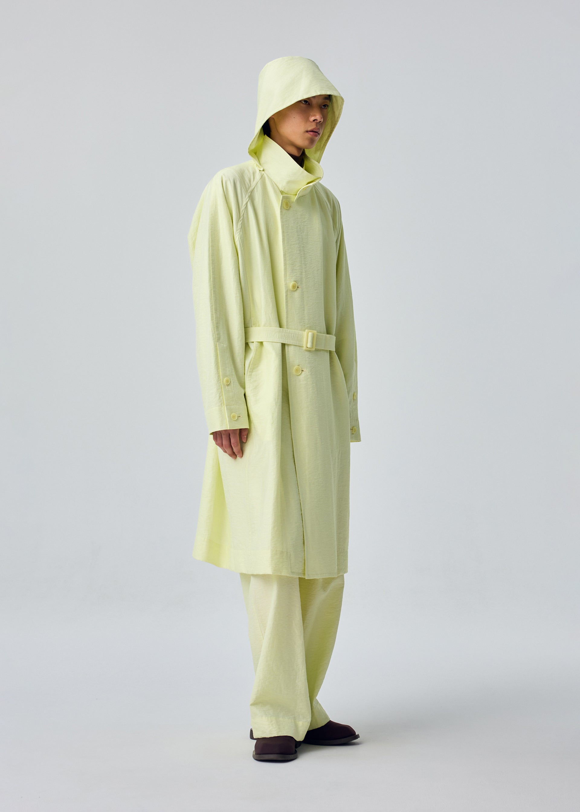 Zoomed view A person stands against a plain white background wearing the OPACITY light yellow hooded coat with detachable hood and matching wide-leg pants, both crafted from a nylon linen blend by HOMME PLISSÉ ISSEY MIYAKE, paired with dark shoes.