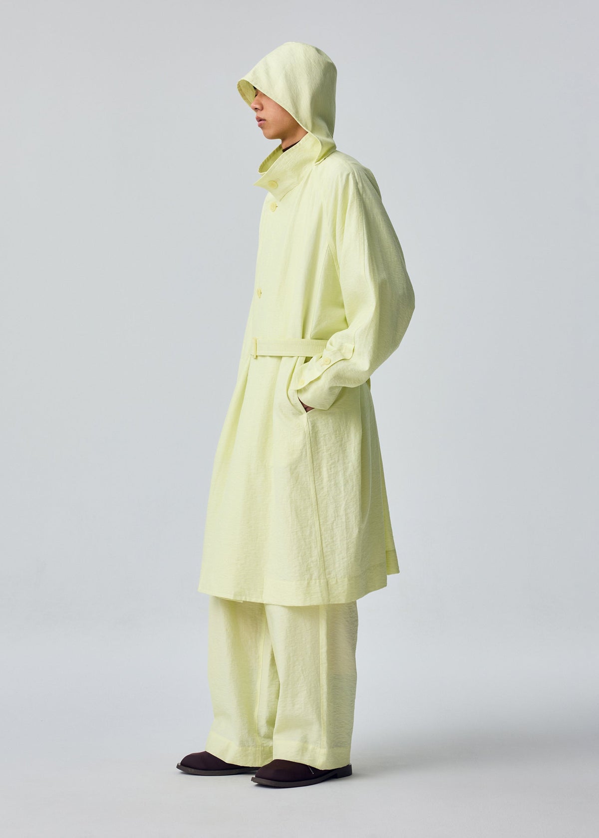 A person stands sideways with hands in pockets against a plain, light background, wearing the OPACITY pale yellow hooded coat with detachable hood and matching wide-leg pants from HOMME PLISSÉ ISSEY MIYAKE. view 9 of 9