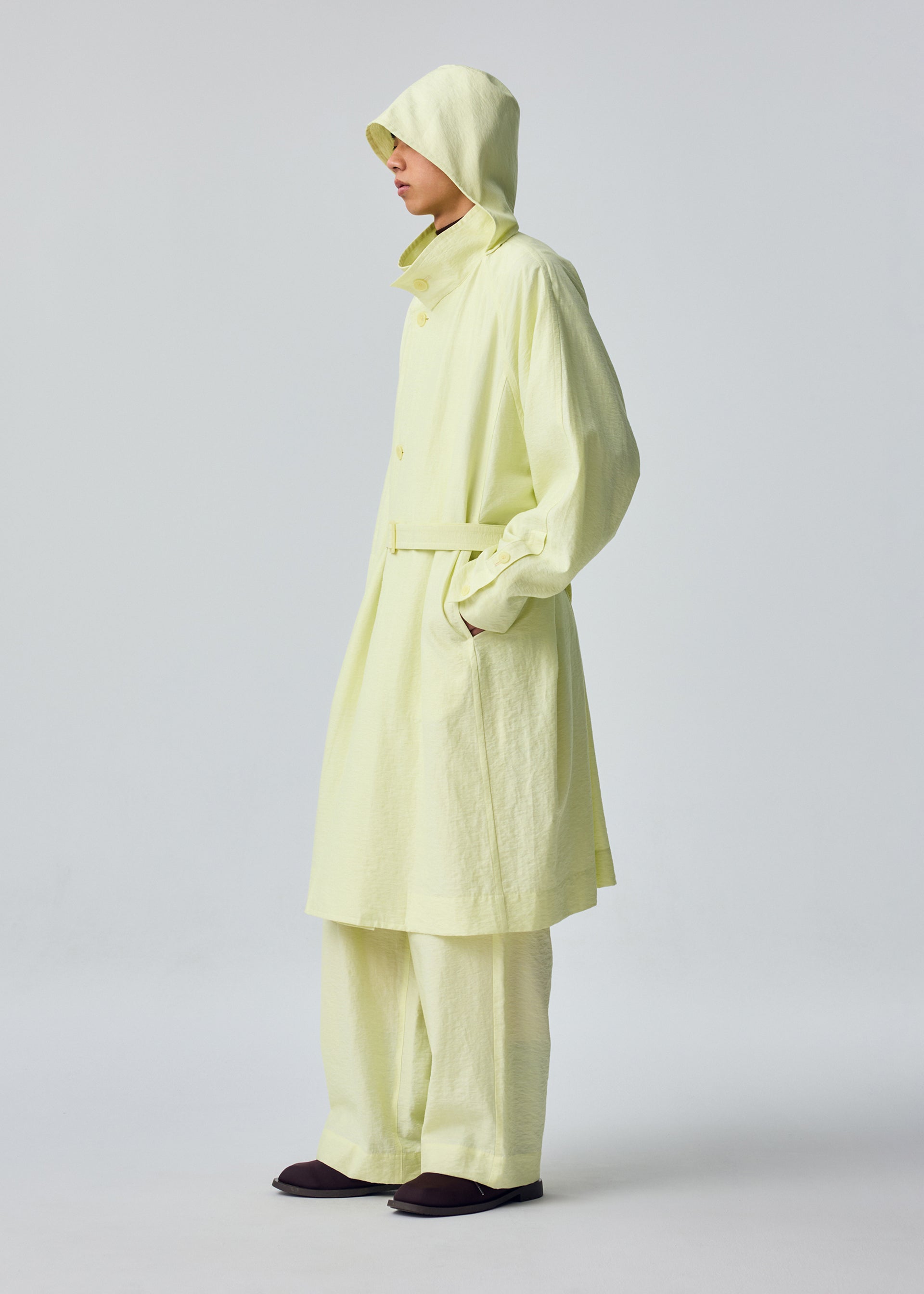 Zoomed view A person stands sideways with hands in pockets against a plain, light background, wearing the OPACITY pale yellow hooded coat with detachable hood and matching wide-leg pants from HOMME PLISSÉ ISSEY MIYAKE.