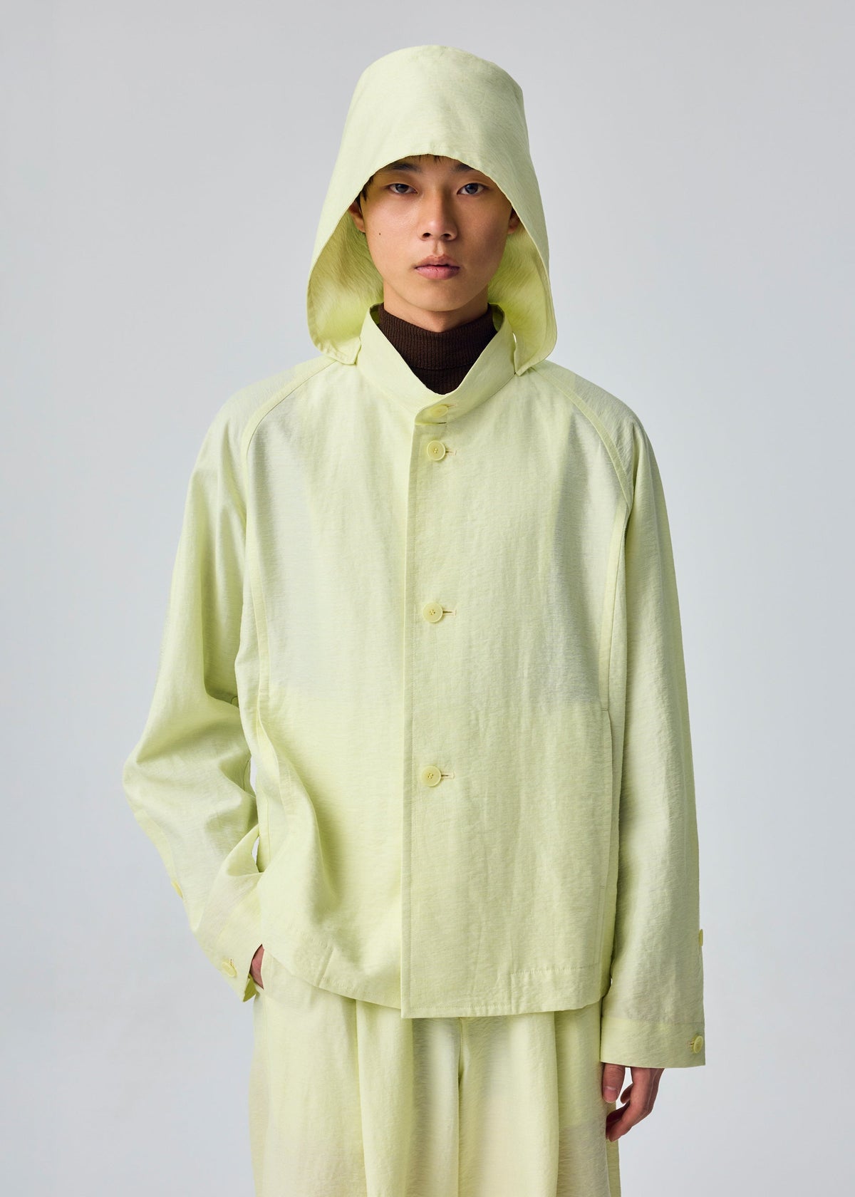 A person wearing the OPACITY light yellow hooded jacket with a detachable hood and matching pants by HOMME PLISSÉ ISSEY MIYAKE stands facing forward against a plain light background. view 6 of 6