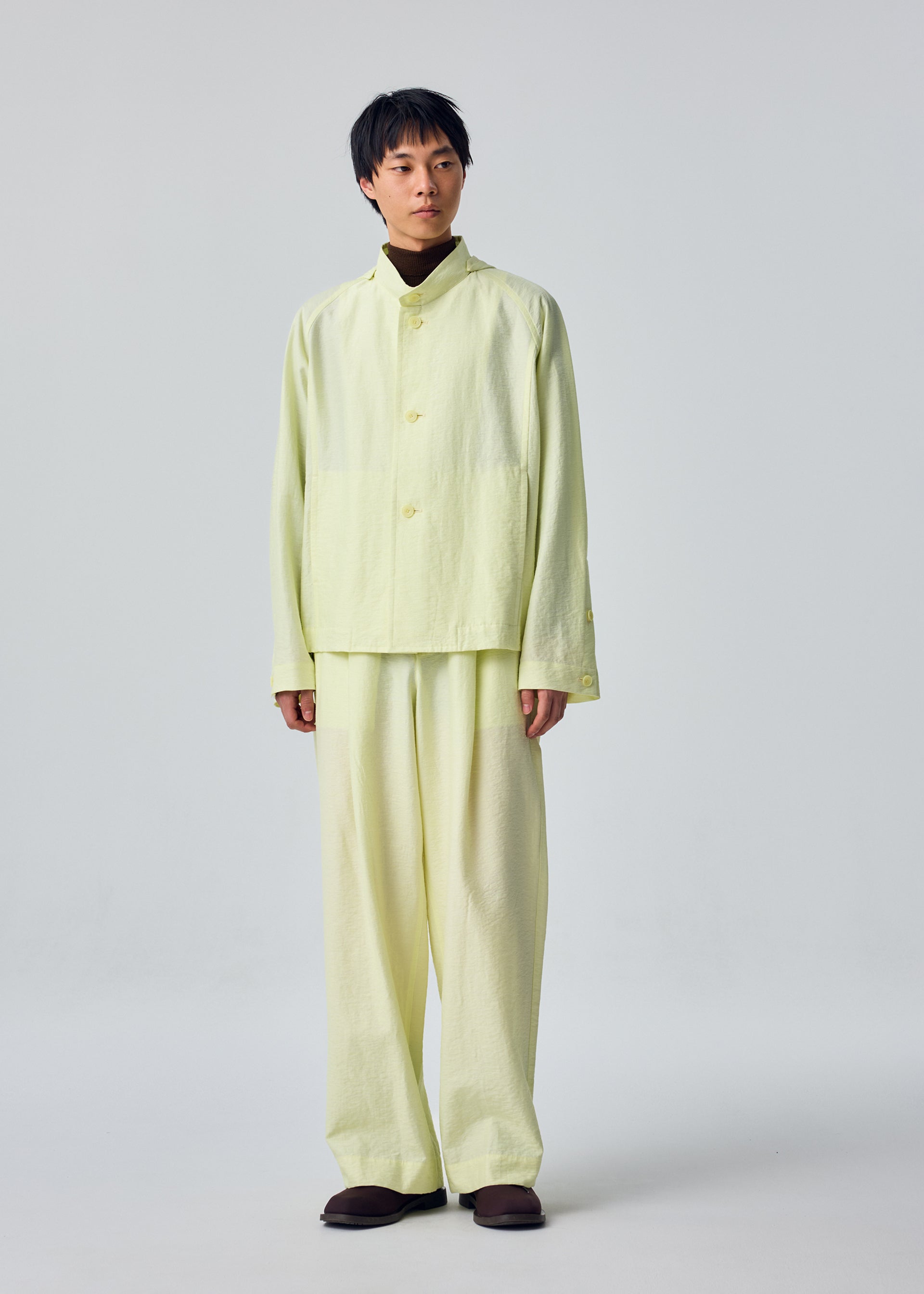 Zoomed view A person stands facing forward, dressed in the OPACITY jacket and matching wide pants from HOMME PLISSÉ ISSEY MIYAKE—a loose-fitting, pale yellow linen-blend ensemble—paired with dark shoes against a plain light background.