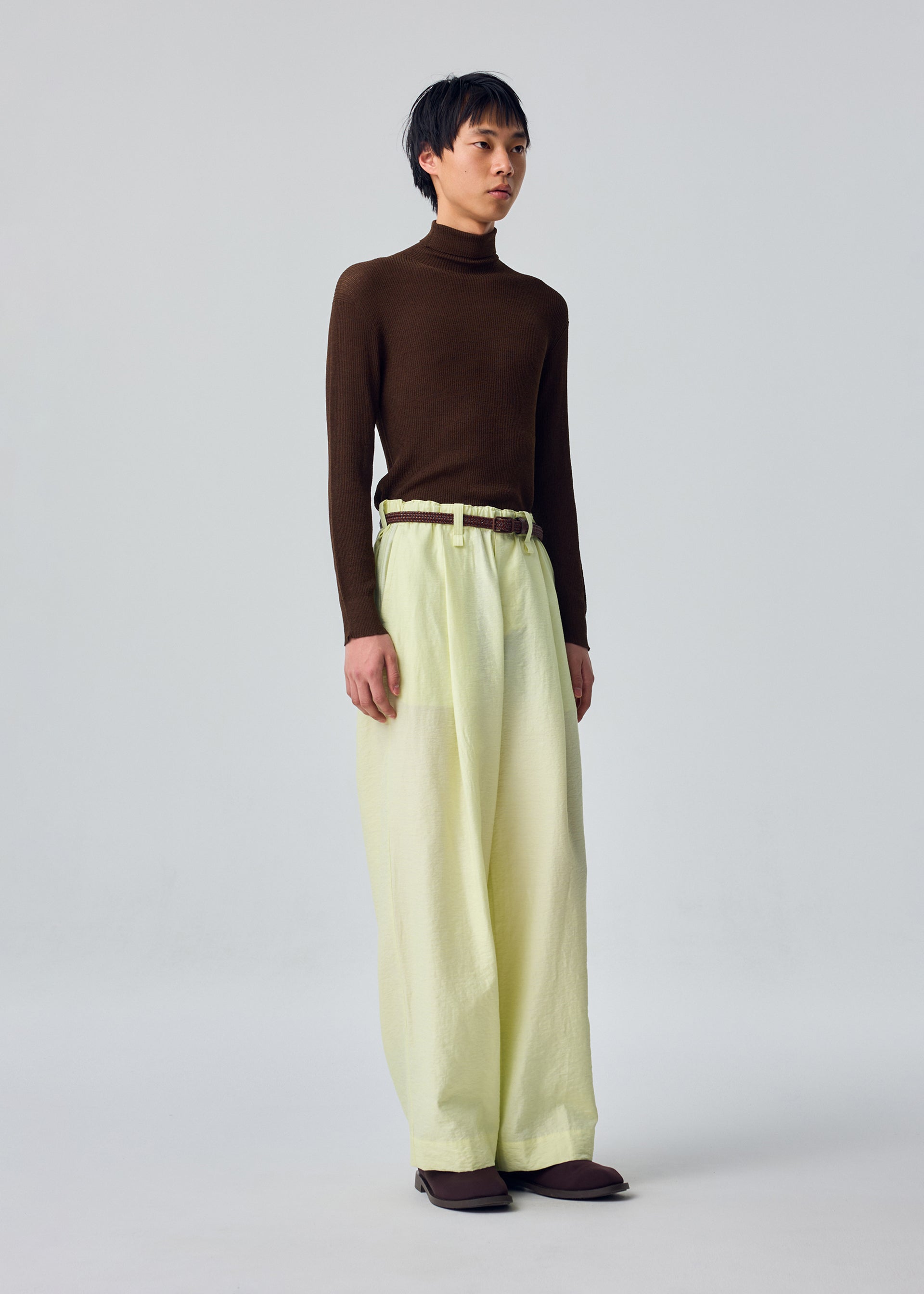 Zoomed view An individual stands against a plain light gray background, dressed in the OPACITY turtleneck by HOMME PLISSÉ ISSEY MIYAKE paired with cream linen blend wide-leg pants and brown shoes.