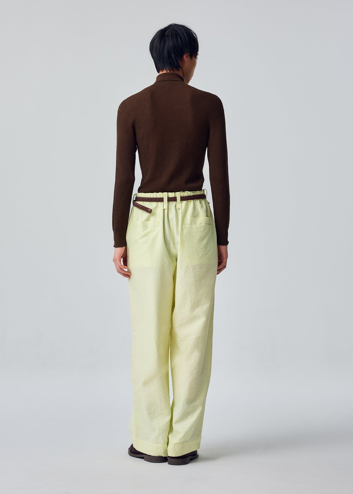 A person stands with their back to the camera, wearing HOMME PLISSÉ ISSEY MIYAKE’s OPACITY wide pants in light yellow—a loose, comfortable style crafted from a linen blend—paired with a brown long-sleeve top and brown belt. The background is plain and light-colored. view 7 of 8