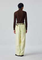A person stands with their back to the camera, wearing HOMME PLISSÉ ISSEY MIYAKE’s OPACITY wide pants in light yellow—a loose, comfortable style crafted from a linen blend—paired with a brown long-sleeve top and brown belt. The background is plain and light-colored.