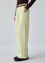 A person stands with one hand in their pocket against a plain background, wearing the OPACITY light yellow linen-blend wide-leg pants from HOMME PLISSÉ ISSEY MIYAKE, styled with a brown belt, a dark brown long-sleeve top, and dark shoes.