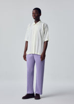 A person stands against a plain light background, wearing the cream-colored EDGE SHIRT by HOMME PLISSÉ ISSEY MIYAKE with vertical stripes and loose, light purple hand-pleated pants, paired with dark brown shoes.