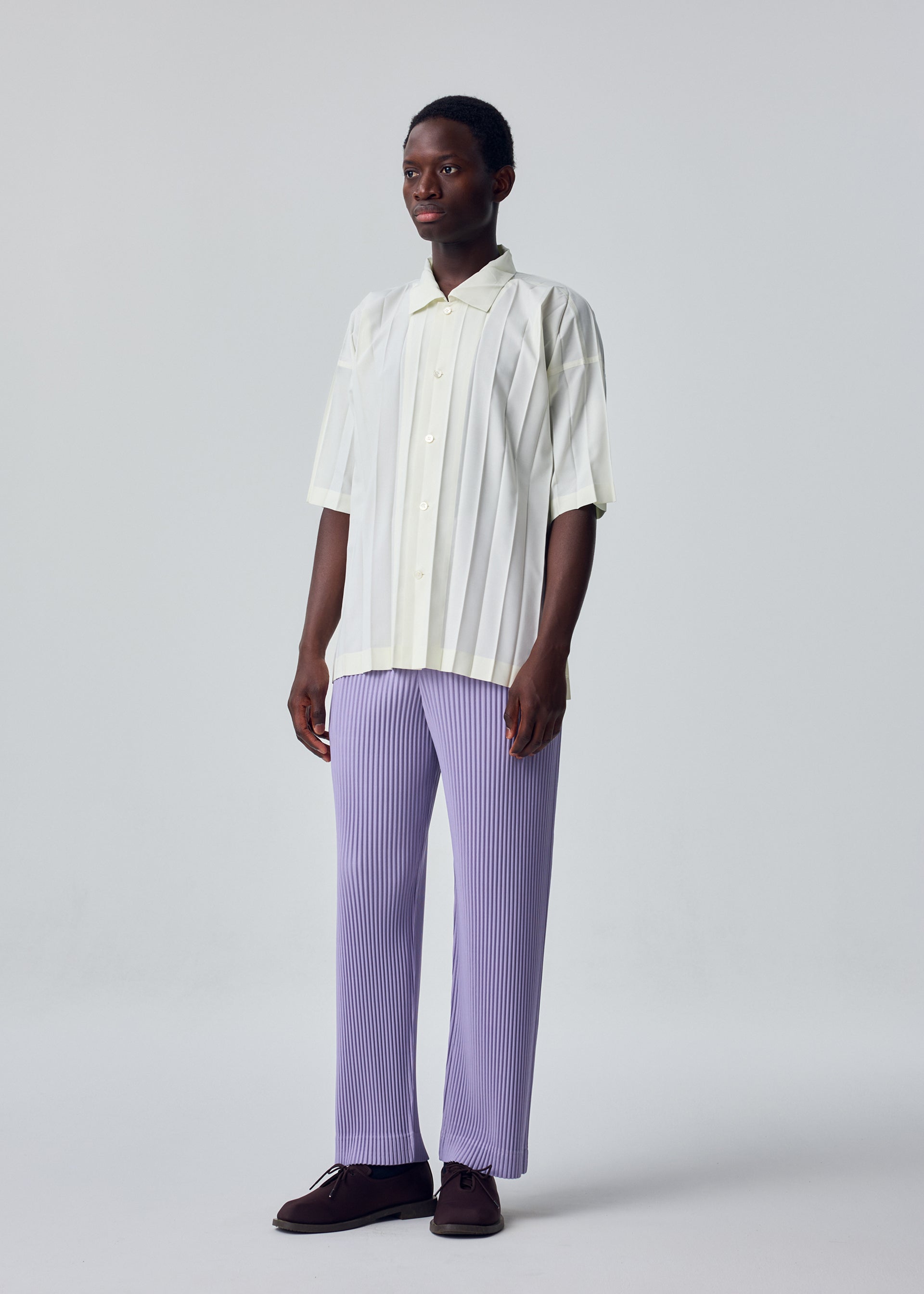 Zoomed view A person stands against a plain light background, wearing the cream-colored EDGE SHIRT by HOMME PLISSÉ ISSEY MIYAKE with vertical stripes and loose, light purple hand-pleated pants, paired with dark brown shoes.
