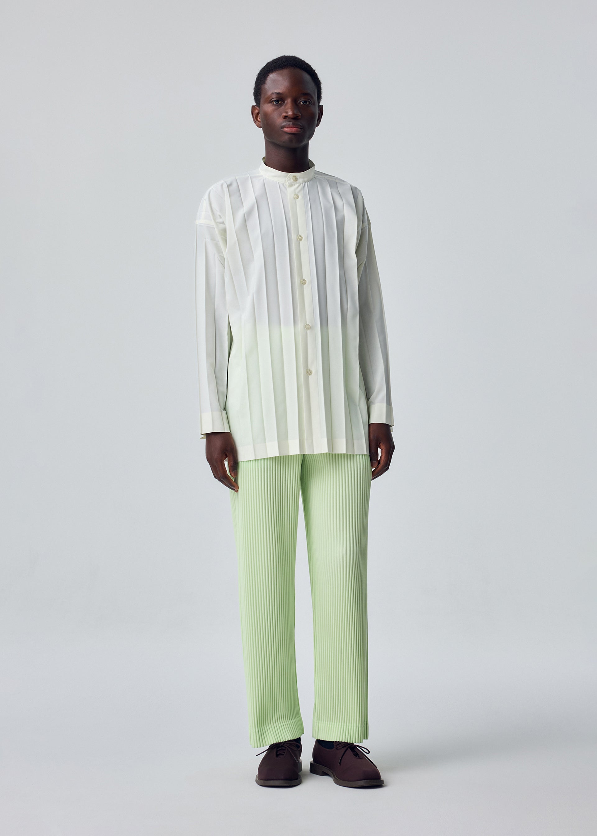 Zoomed view A person stands against a plain background wearing the relaxed fit, white EDGE SHIRT by HOMME PLISSÉ ISSEY MIYAKE with hand pleating and light green pleated pants, paired with dark brown shoes.