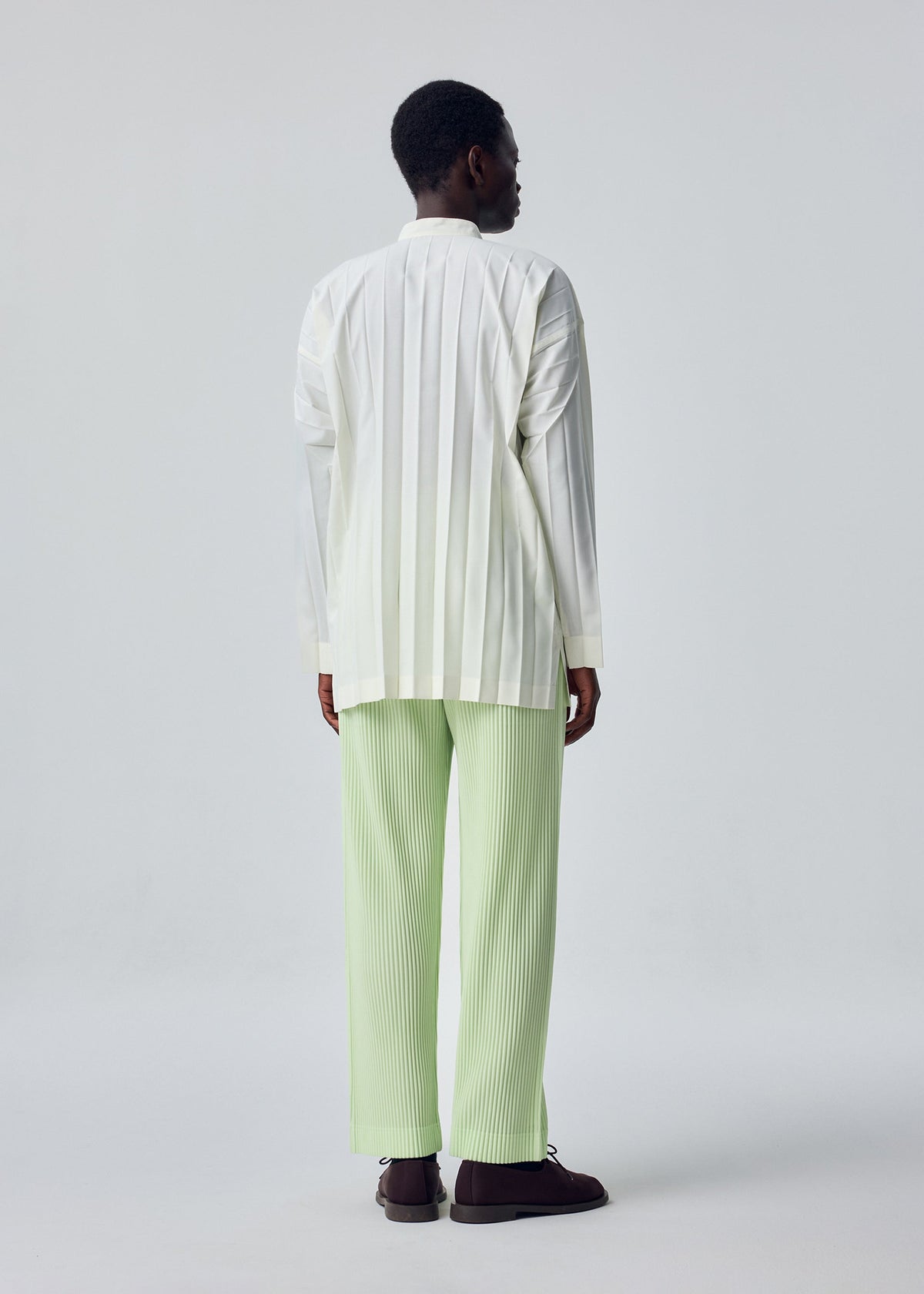 A person with short hair stands against a plain light background, wearing the HOMME PLISSÉ ISSEY MIYAKE EDGE SHIRT with hand pleating and relaxed-fit light green pleated pants, finished with dark shoes. view 7 of 7