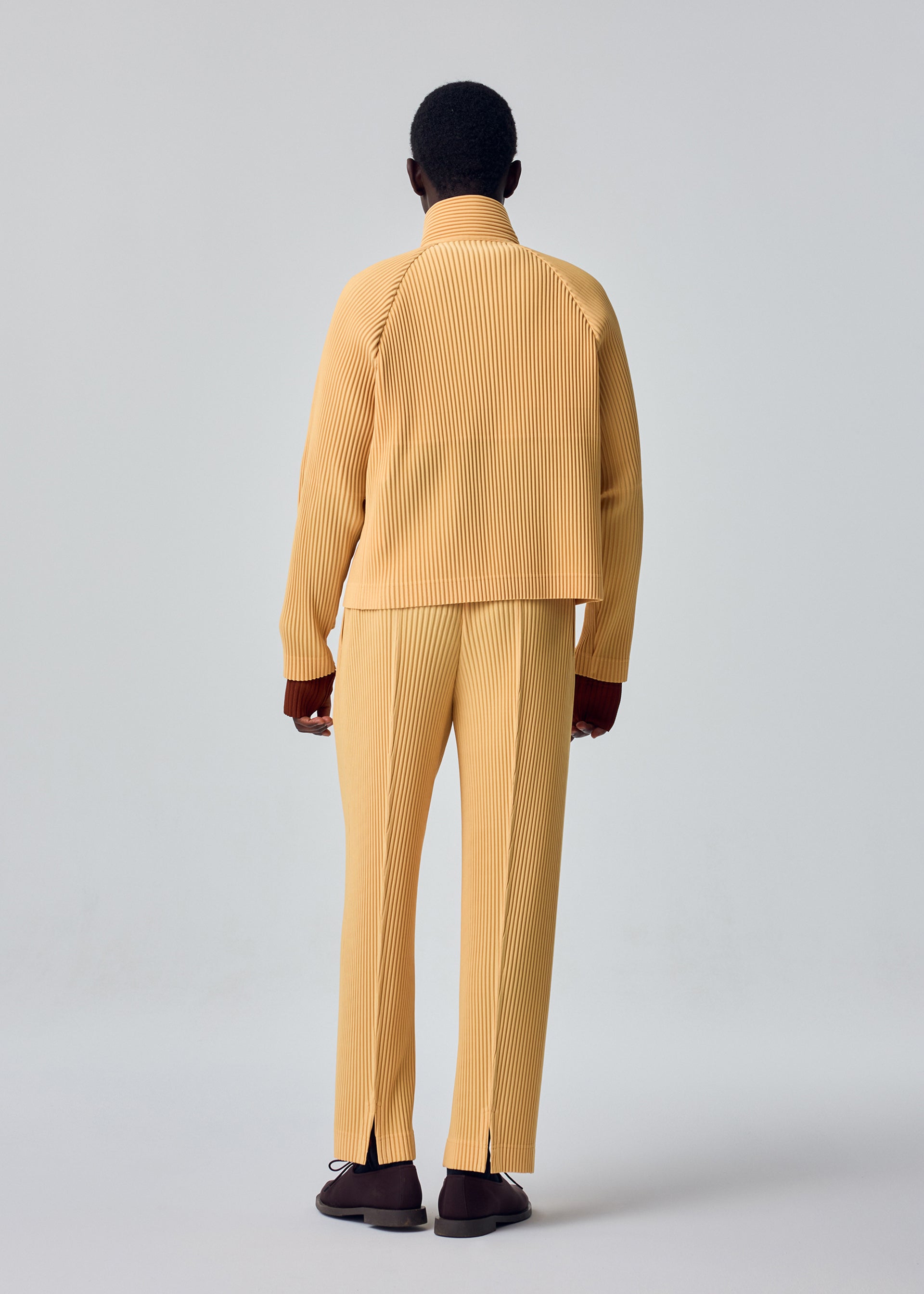 Zoomed view A person stands facing away, dressed in the HOMME PLISSÉ ISSEY MIYAKE TAILORED PLEATS 1 set—a matching light yellow, ribbed jacket and pants with subtle diagonal pleats—paired with brown shoes, against a plain light gray background.