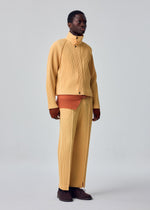 A person stands against a plain light gray background wearing the TAILORED PLEATS 1 jacket and pants from HOMME PLISSÉ ISSEY MIYAKE in beige, layered over a rust-orange pleated top and paired with brown shoes. The ensemble features subtle diagonal pleats and is crafted from quick-drying polyester, combining style with practicality.