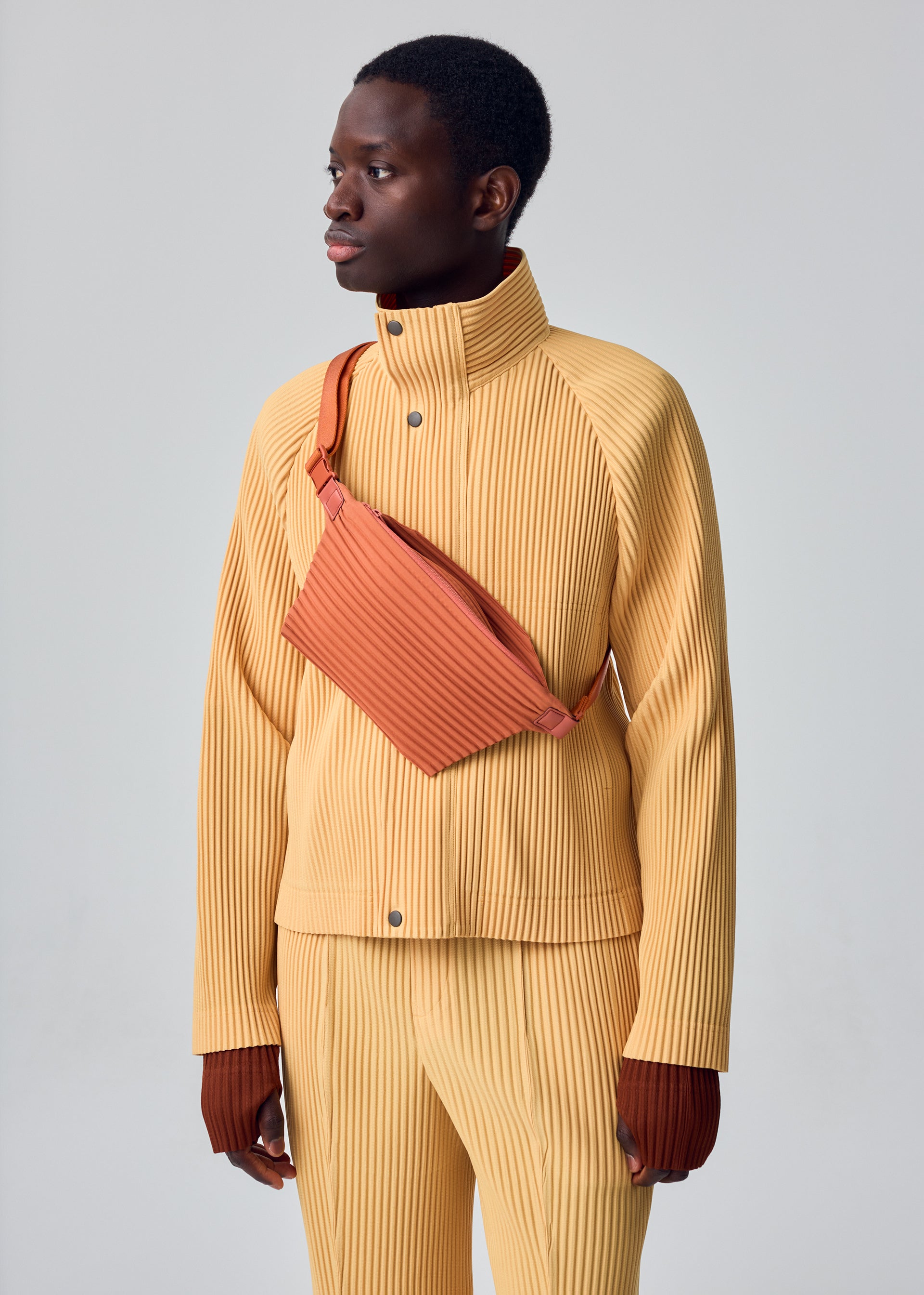 Zoomed view A person stands against a plain light gray background, wearing the TAILORED PLEATS 1 jacket and pants set by HOMME PLISSÉ ISSEY MIYAKE in yellow. The ribbed raglan ensemble features a high collar, silver snaps, and diagonal pleats, crafted from quick-drying polyester. They accessorize with a rust-colored crossbody bag and brown gloves.