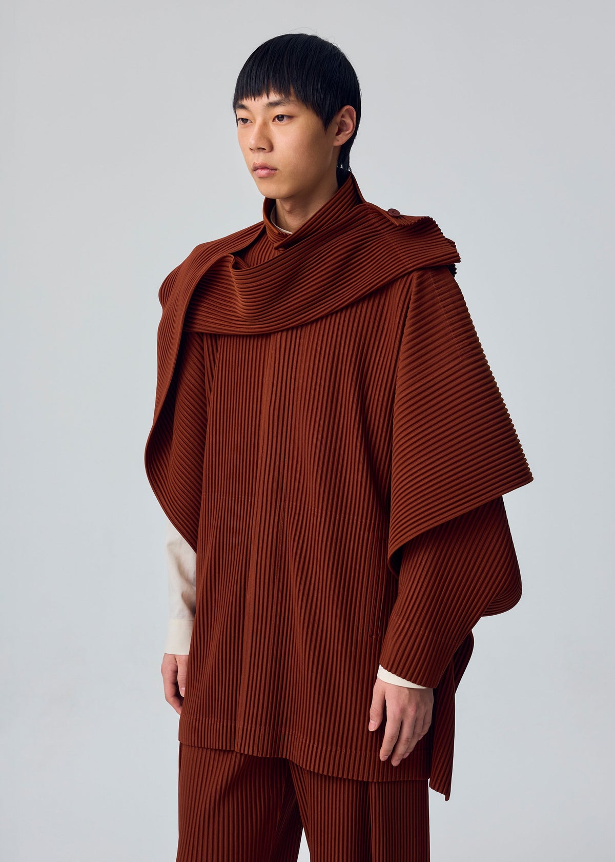 A person with short black hair wears the MOTION BLUR pleated jersey jacket by HOMME PLISSÉ ISSEY MIYAKE in rust, styled as a coordinated outfit against a plain light gray background. view 4 of 5