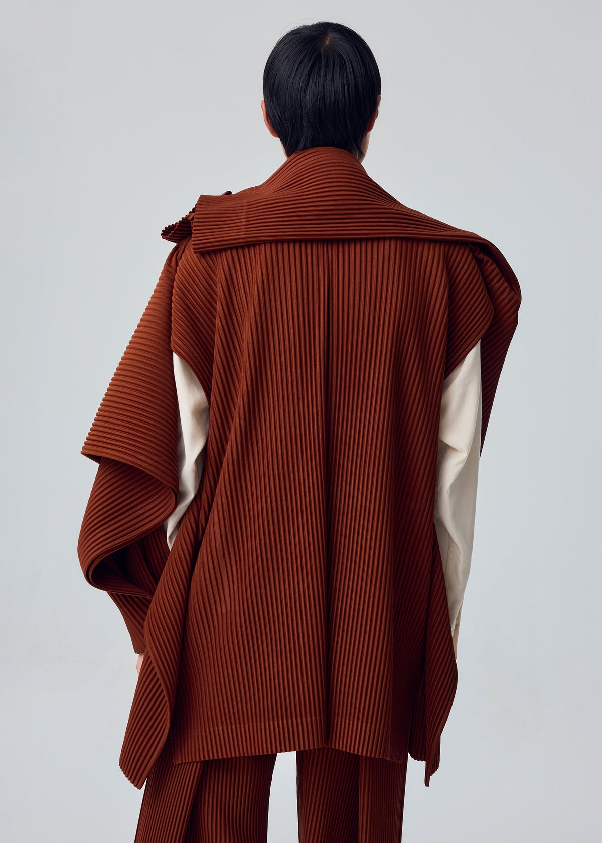A person stands with their back to the camera, wearing the MOTION BLUR brown pleated jersey jacket with cape-like detail over cream sleeves by HOMME PLISSÉ ISSEY MIYAKE, set against a plain light background. view 5 of 5