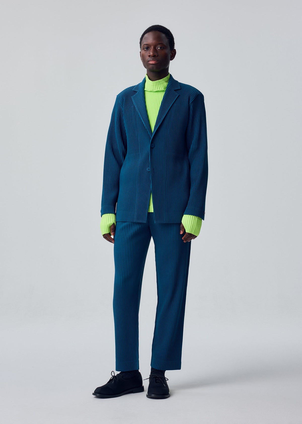 A person stands against a plain light background, dressed in the HOMME PLISSÉ ISSEY MIYAKE TAILORED PLEATS 1—a loose blue pinstripe suit with a formal-casual blazer—layered over a bright neon green turtleneck sweater and paired with black shoes. view 5 of 6