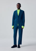A person stands against a plain light background, dressed in the HOMME PLISSÉ ISSEY MIYAKE TAILORED PLEATS 1—a loose blue pinstripe suit with a formal-casual blazer—layered over a bright neon green turtleneck sweater and paired with black shoes.