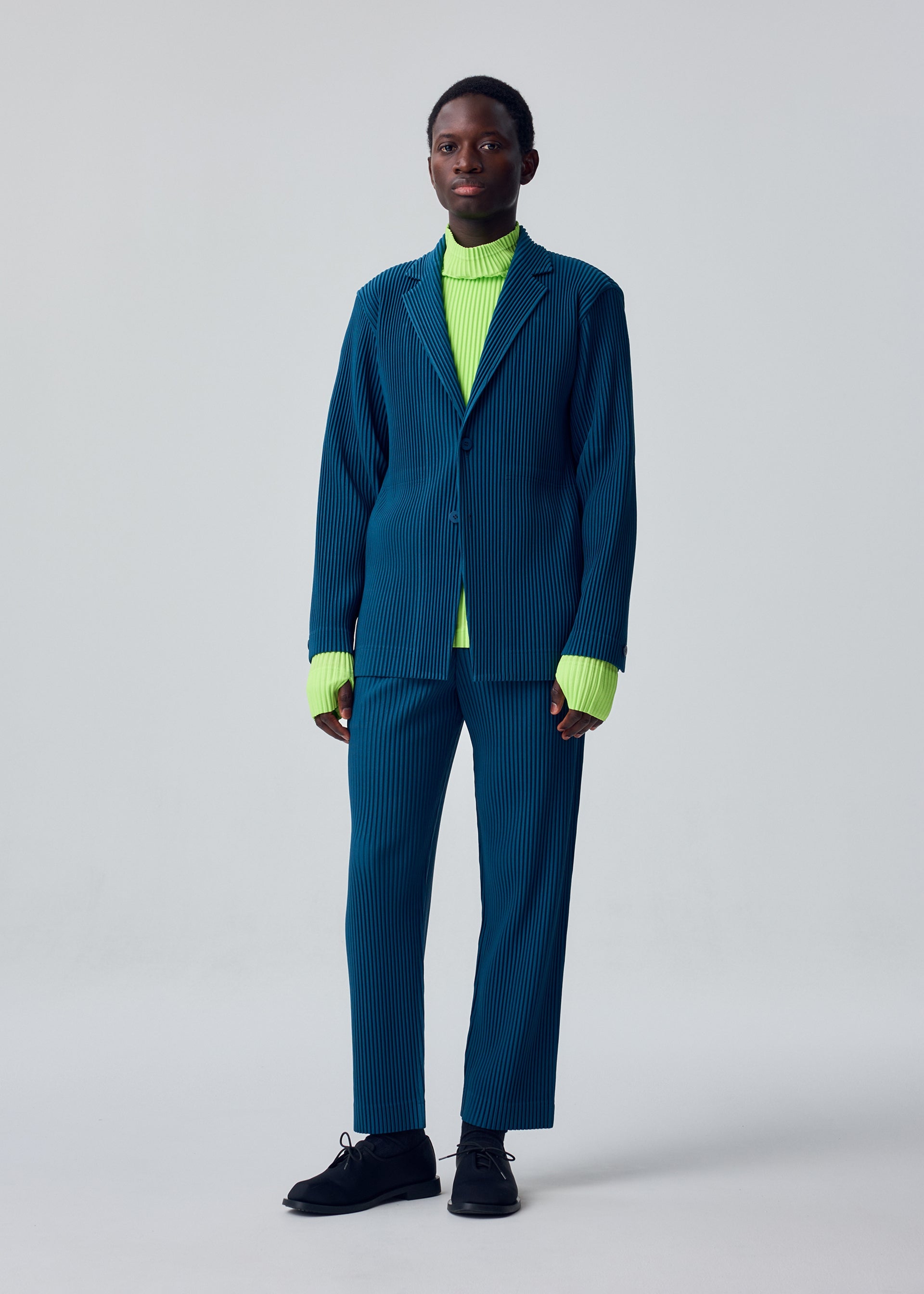 Zoomed view A person stands against a plain light background, dressed in the HOMME PLISSÉ ISSEY MIYAKE TAILORED PLEATS 1—a loose blue pinstripe suit with a formal-casual blazer—layered over a bright neon green turtleneck sweater and paired with black shoes.