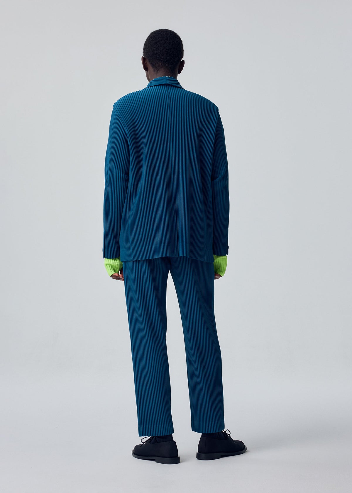 A person stands facing away, dressed in the HOMME PLISSÉ ISSEY MIYAKE TAILORED PLEATS 1—a blue pinstriped suit with wide-leg, cuffed trousers and a sharply tailored jacket. Neon green sleeves emerge from beneath the blazer, set against a plain light gray background. view 6 of 6
