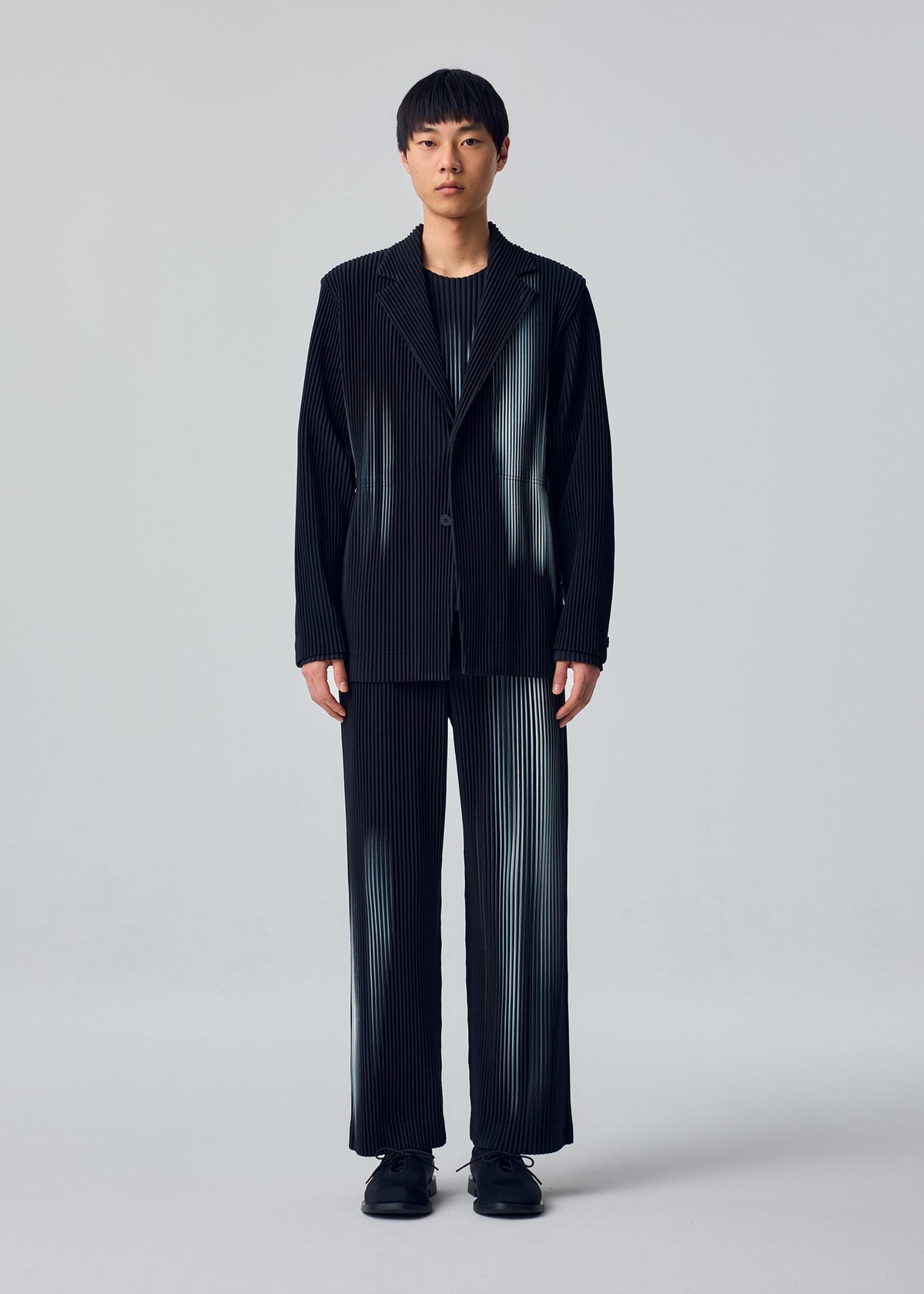 Zoomed view A person stands facing forward wearing the IN THE SHADOW jacket by HOMME PLISSÉ ISSEY MIYAKE, paired with a black shirt and shoes, against a plain light gray background.