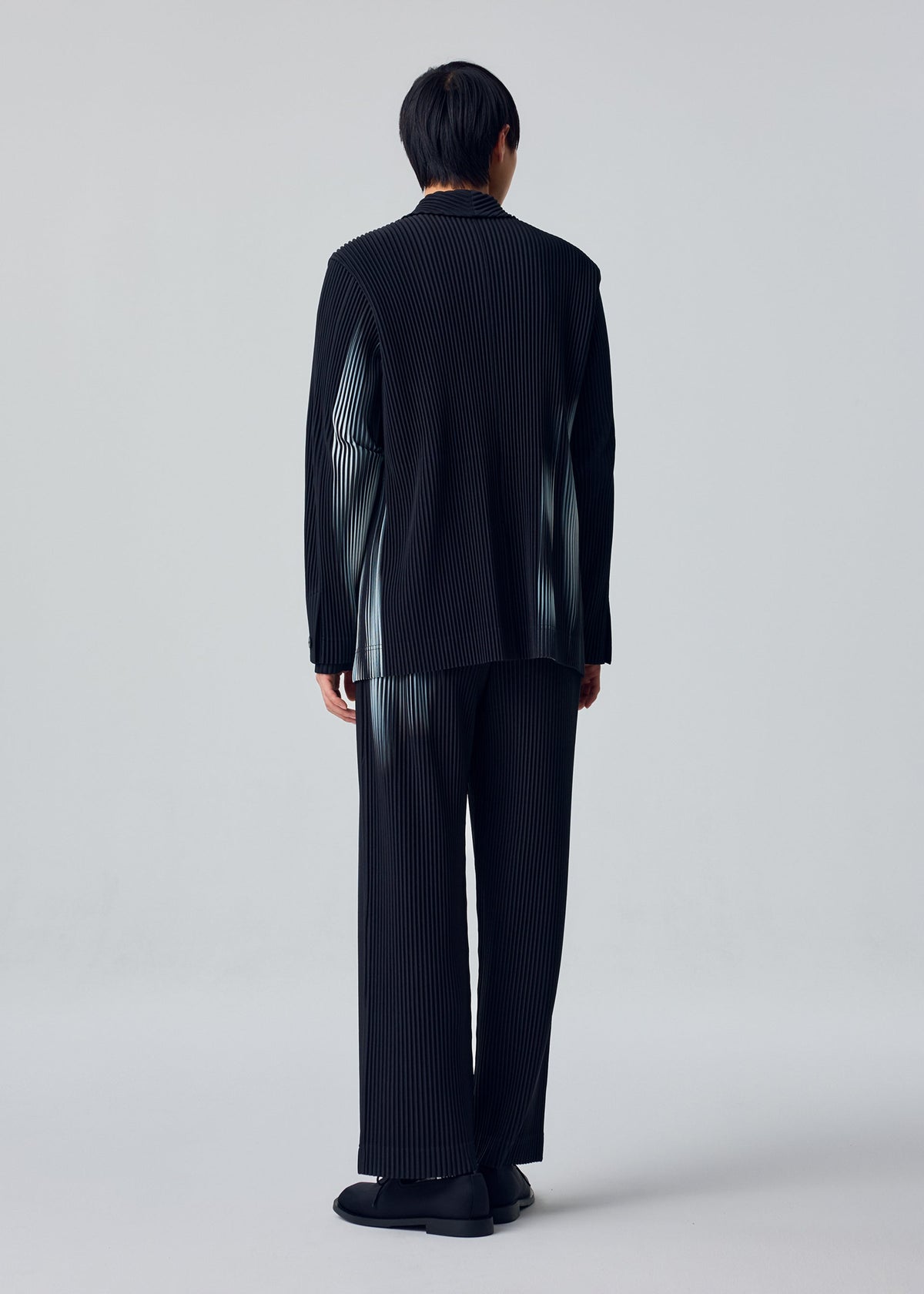 Zoomed view A person faces away wearing the IN THE SHADOW black pinstripe suit by HOMME PLISSÉ ISSEY MIYAKE, with matching pants and black shoes against a plain light background.