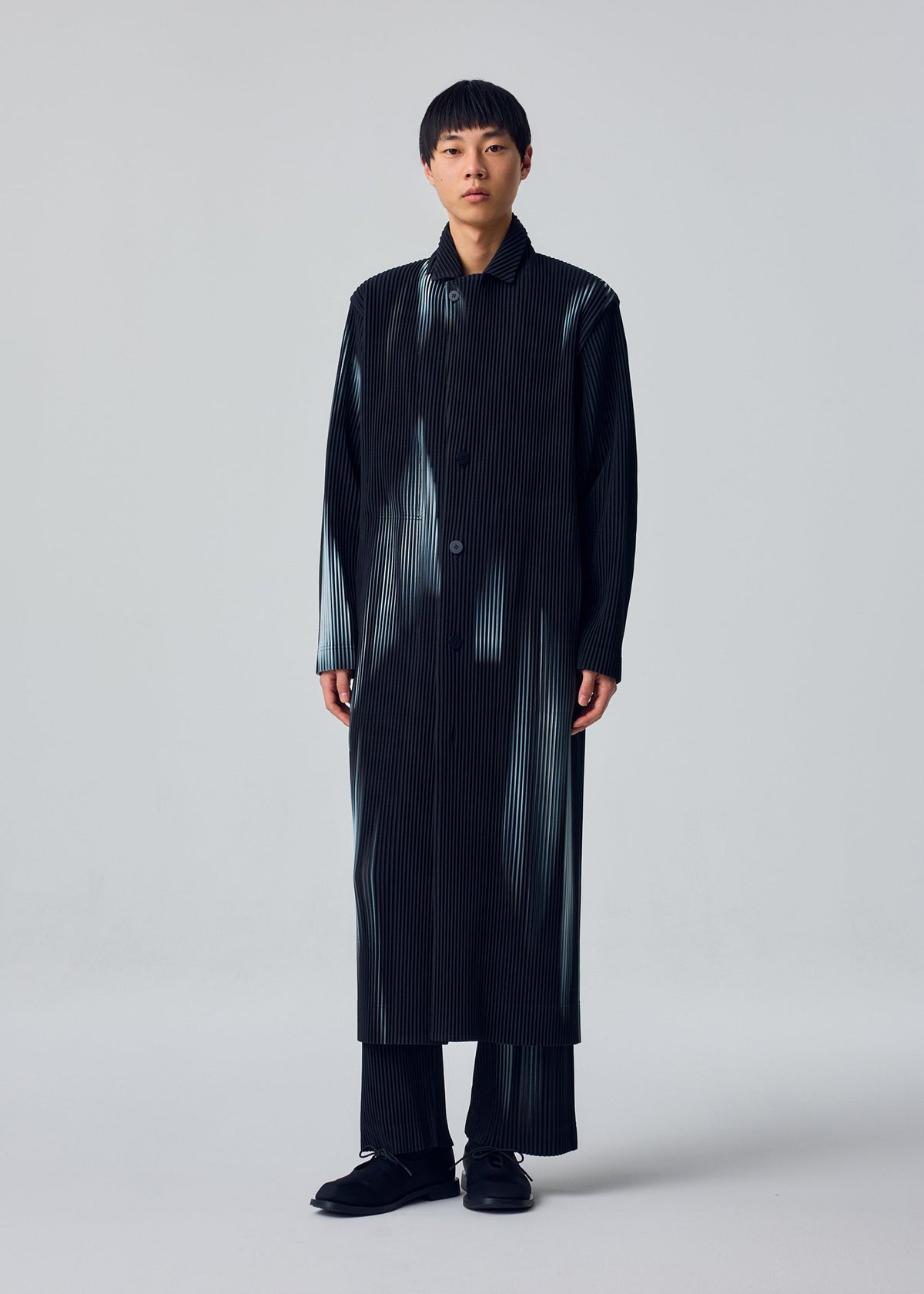 Zoomed view A person with short hair and a neutral expression wears the IN THE SHADOW jacket by HOMME PLISSÉ ISSEY MIYAKE over matching pinstriped pants and black shoes, standing against a plain background. The jacket is loose and buttoned up front.