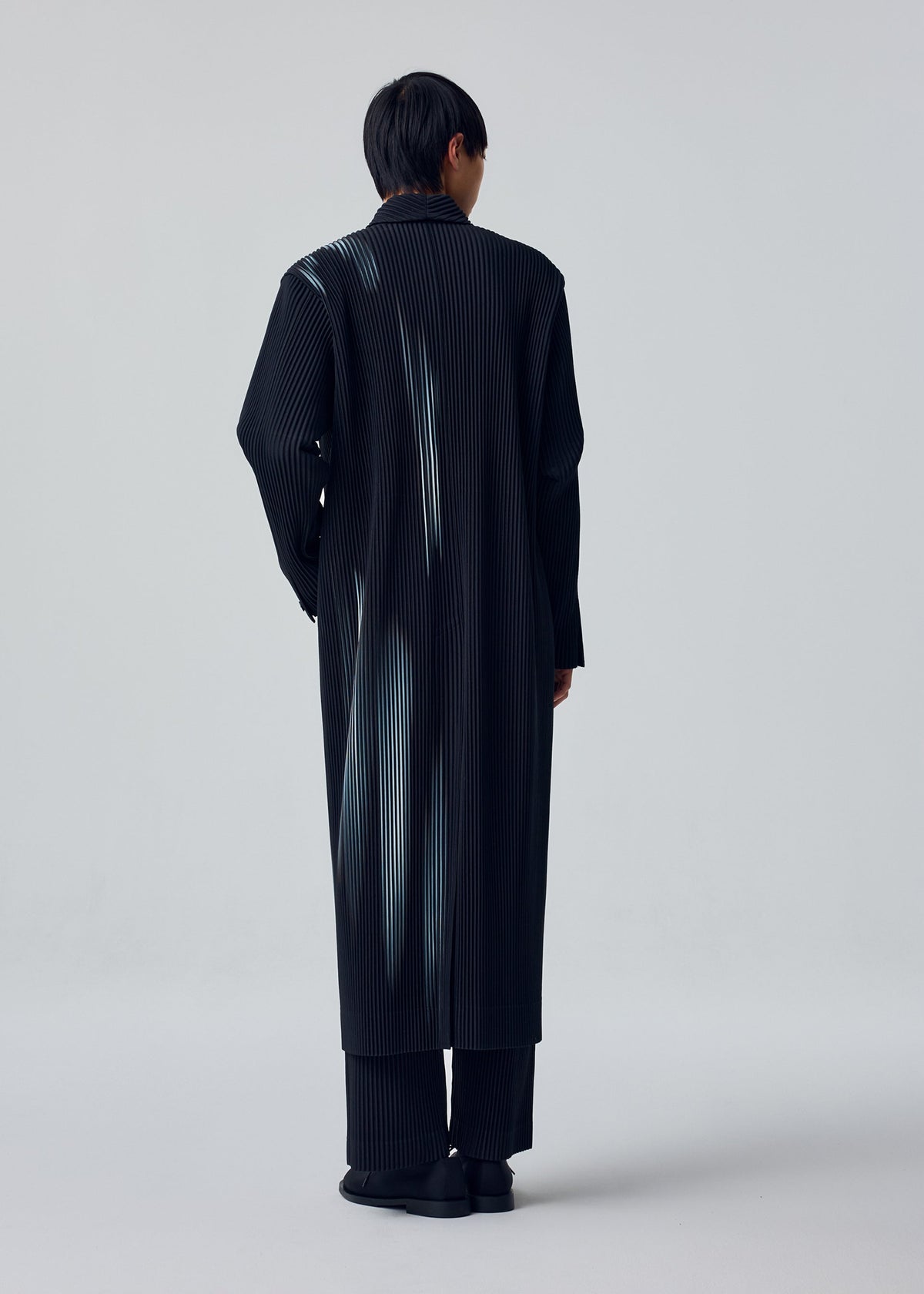 A person with short dark hair models the IN THE SHADOW by HOMME PLISSÉ ISSEY MIYAKE: a long, black, optical-stripe coat over matching pants and black shoes in a minimalist studio with a light gray backdrop. view 5 of 5