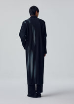 A person with short dark hair models the IN THE SHADOW by HOMME PLISSÉ ISSEY MIYAKE: a long, black, optical-stripe coat over matching pants and black shoes in a minimalist studio with a light gray backdrop.