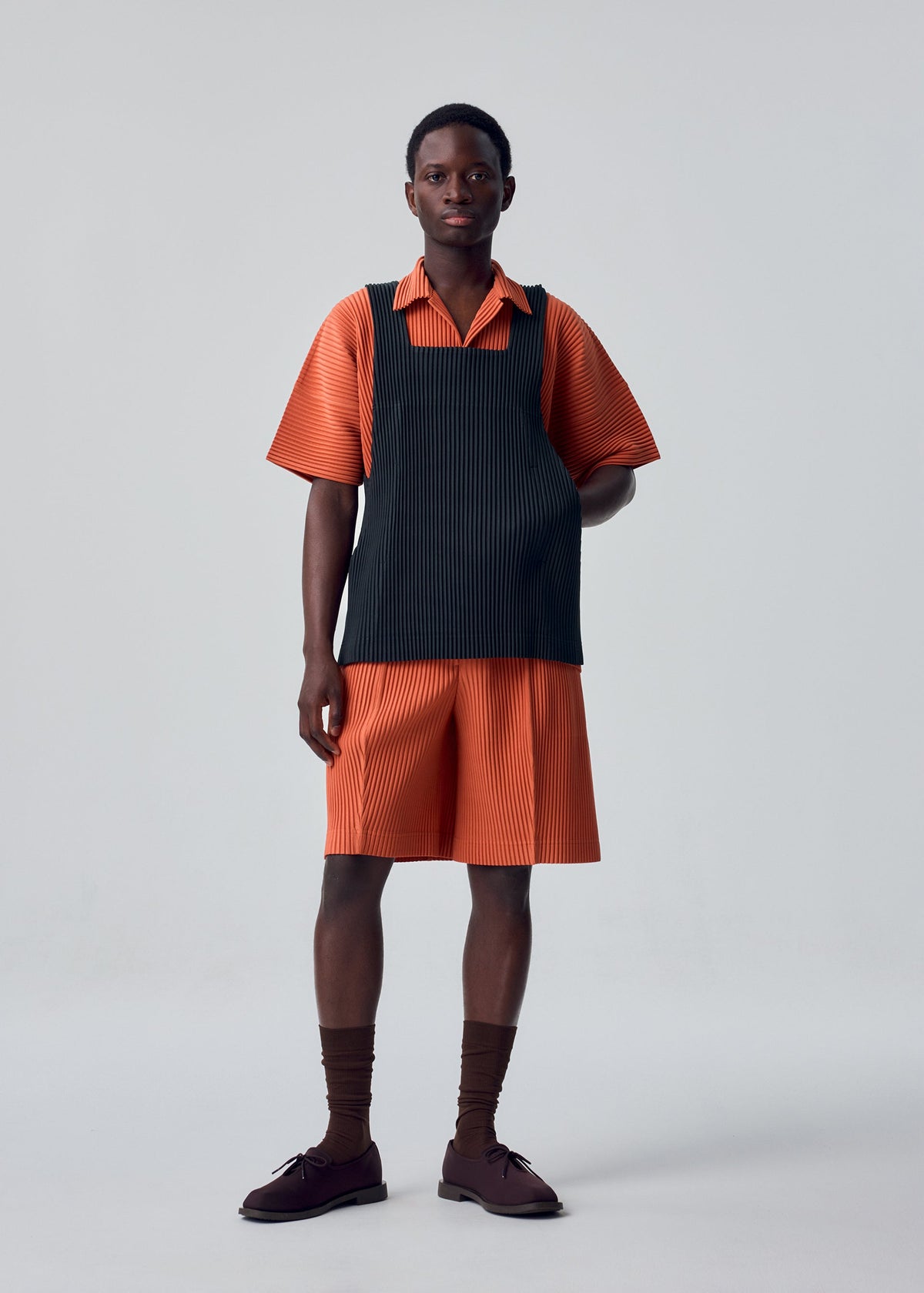 A person stands in a studio against a plain light gray background, wearing items from the HOMME PLISSÉ ISSEY MIYAKE MONTHLY COLORS : AUGUST collection: an orange short-sleeve shirt and matching shorts, paired with a black apron-style vest crafted from quick-drying polyester for lightweight comfort, completed with brown socks and brown shoes. view 5 of 8