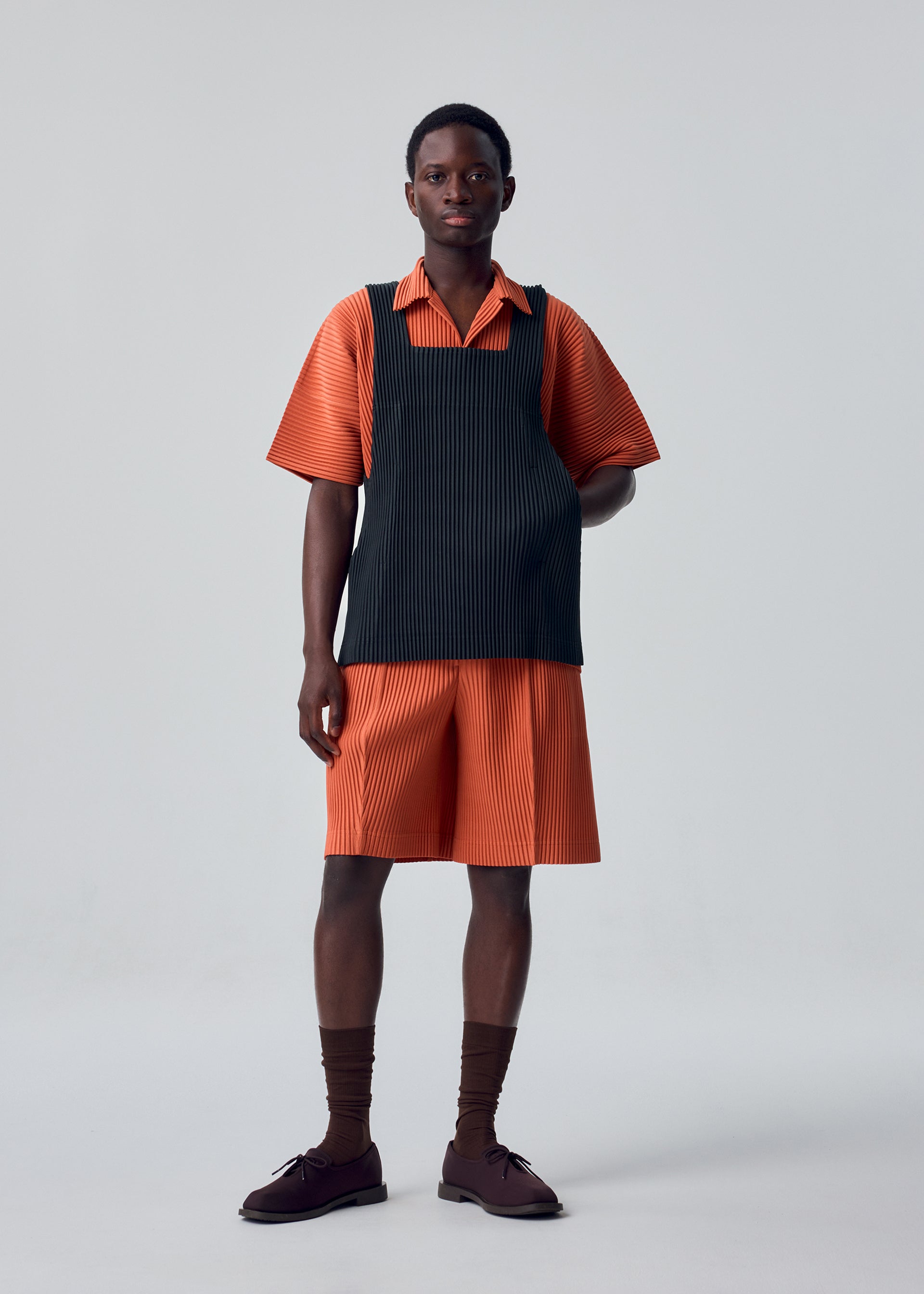 Zoomed view A person stands in a studio against a plain light gray background, wearing items from the HOMME PLISSÉ ISSEY MIYAKE MONTHLY COLORS : AUGUST collection: an orange short-sleeve shirt and matching shorts, paired with a black apron-style vest crafted from quick-drying polyester for lightweight comfort, completed with brown socks and brown shoes.