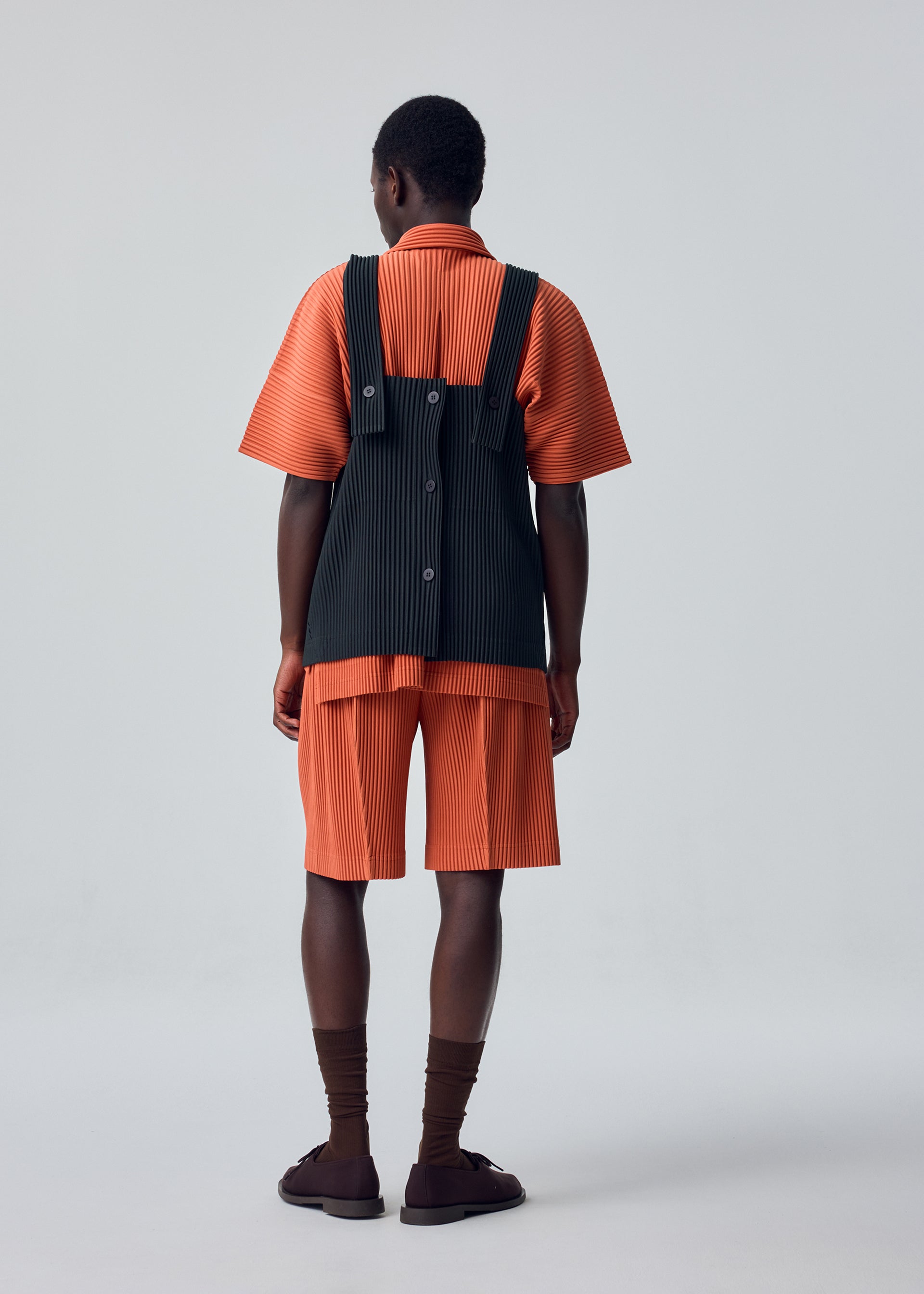 Zoomed view A person stands facing away, dressed in the HOMME PLISSÉ ISSEY MIYAKE “MONTHLY COLORS : AUGUST” set—an orange pleated shirt and shorts made from quick-drying polyester for lightweight comfort. The look is layered with a black, apron-style pinstriped vest adorned with large buttons, and completed with brown socks and sandals against a plain background.