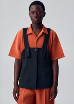 A person stands against a plain background wearing the MONTHLY COLORS : AUGUST set from HOMME PLISSÉ ISSEY MIYAKE—an ensemble featuring an orange short-sleeve shirt and matching orange shorts, layered with a lightweight, button-fastened apron-style vest for added comfort.
