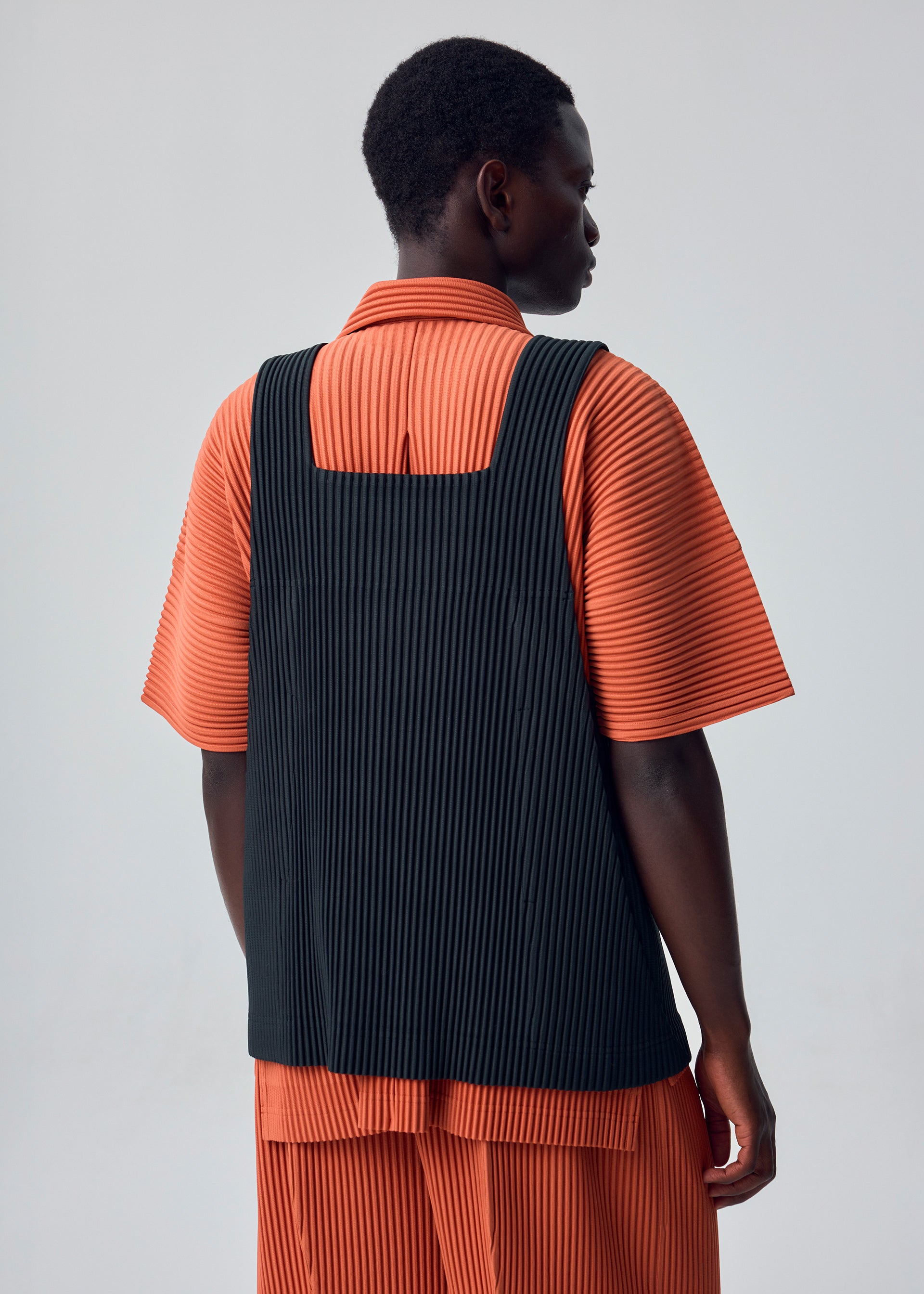 Zoomed view A person stands with their back to the camera against a plain light gray background, wearing the HOMME PLISSÉ ISSEY MIYAKE MONTHLY COLORS : AUGUST set—an orange, pleated short-sleeve shirt and shorts—layered with a black, pleated apron-style vest.