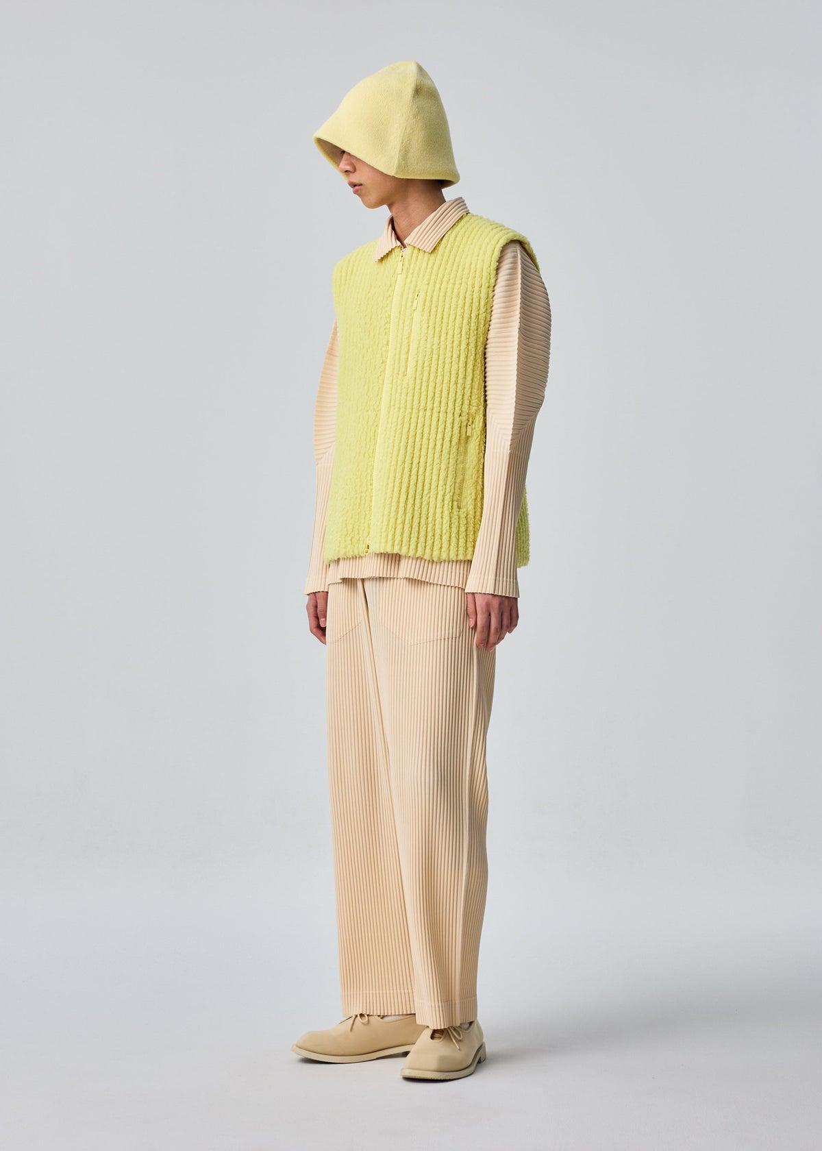 A person in HOMME PLISSÉ ISSEY MIYAKE FLEECE PLEATS, styled with a yellow zip-up vest, cream pleated shirt and pants, matching yellow hat, and cream shoes, stands before a plain light grey background. view 6 of 7
