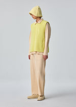 A person in HOMME PLISSÉ ISSEY MIYAKE FLEECE PLEATS, styled with a yellow zip-up vest, cream pleated shirt and pants, matching yellow hat, and cream shoes, stands before a plain light grey background.