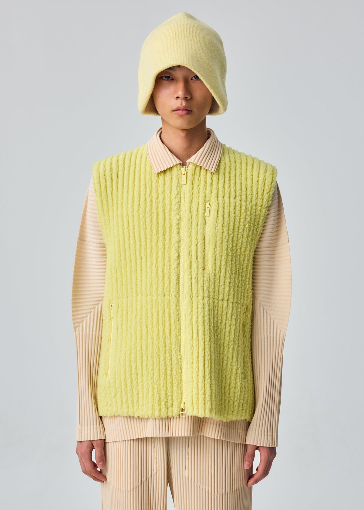 Zoomed view A person in the HOMME PLISSÉ ISSEY MIYAKE FLEECE PLEATS vest, matching hat, and beige pleated shirt and pants stands facing the camera against a plain light background.
