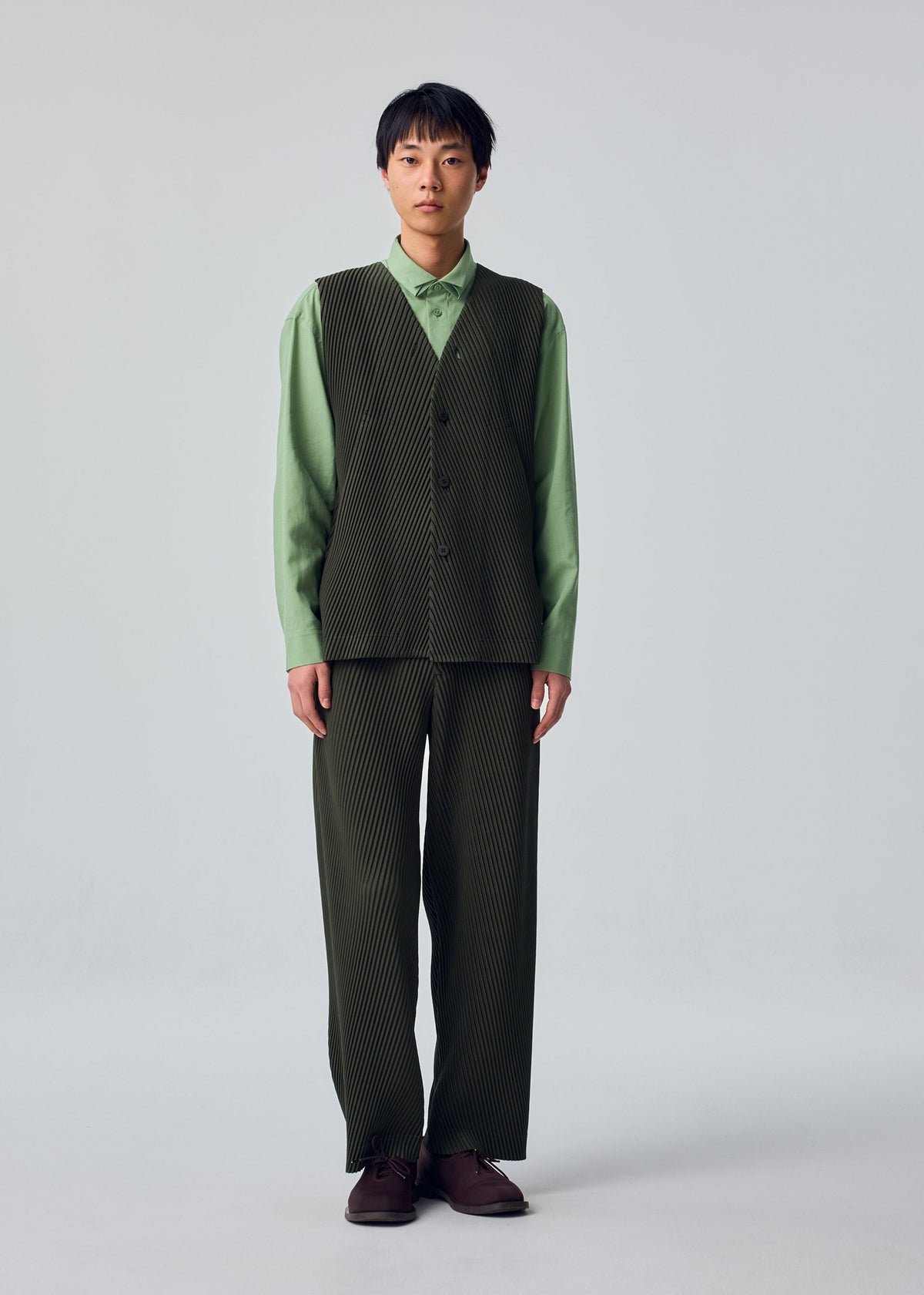 A person stands facing forward, wearing the HOMME PLISSÉ ISSEY MIYAKE SLASH set: a light green collared shirt underneath a dark green, front-opening pinstriped vest and matching pinstriped pants with slash pockets, paired with brown shoes, against a plain light background. view 6 of 8