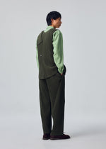 A person stands facing away, wearing the green SLASH shirt and a dark pinstriped SLASH suit with loose pants featuring slash pockets from HOMME PLISSÉ ISSEY MIYAKE, hands in pockets, against a plain light gray background.