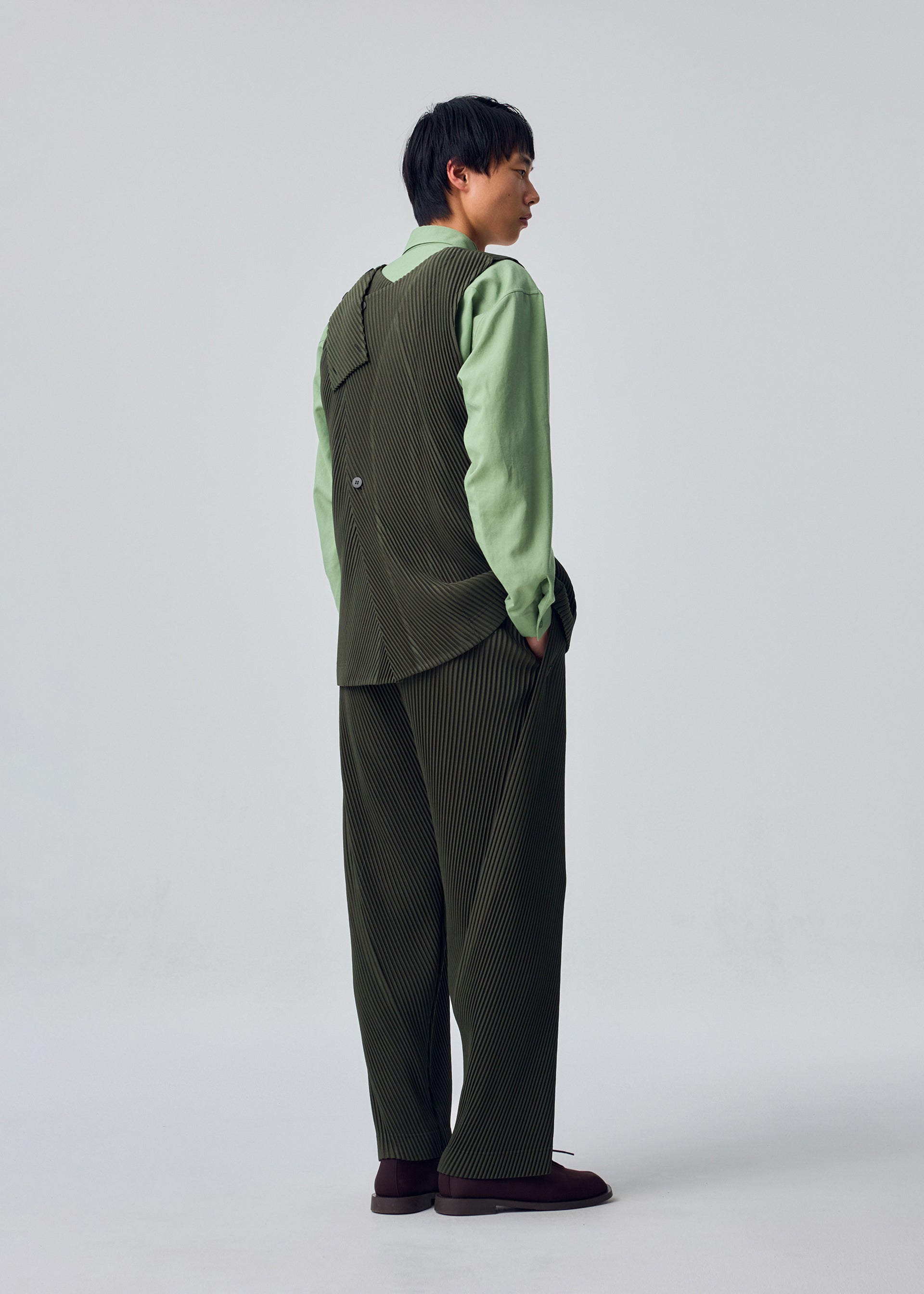 Zoomed view A person stands facing away, wearing the green SLASH shirt and a dark pinstriped SLASH suit with loose pants featuring slash pockets from HOMME PLISSÉ ISSEY MIYAKE, hands in pockets, against a plain light gray background.