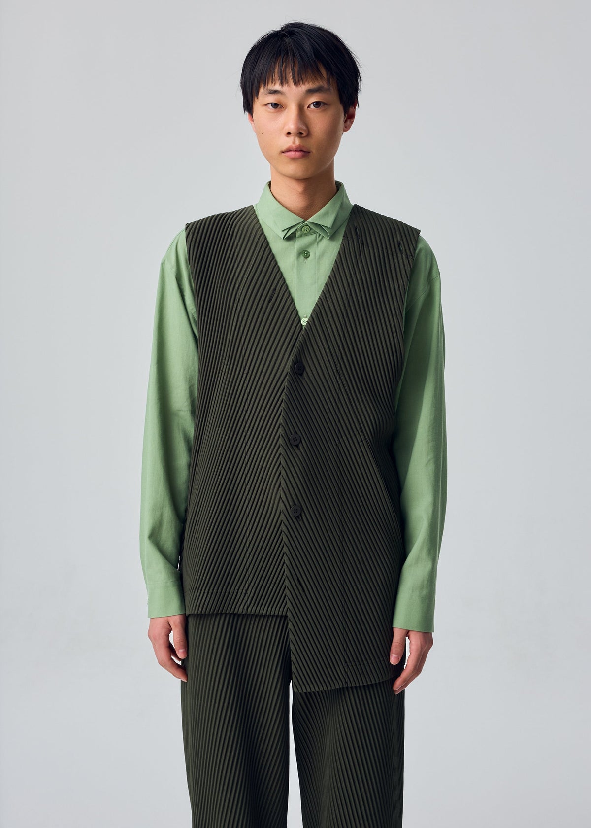 A model wears HOMME PLISSÉ ISSEY MIYAKE’s SLASH ensemble: a light green shirt paired with a dark green, pinstriped vest and matching pants featuring subtle diagonal pleats and slash pockets. Posed against a plain light background, the model gazes at the camera with a neutral expression. view 8 of 8